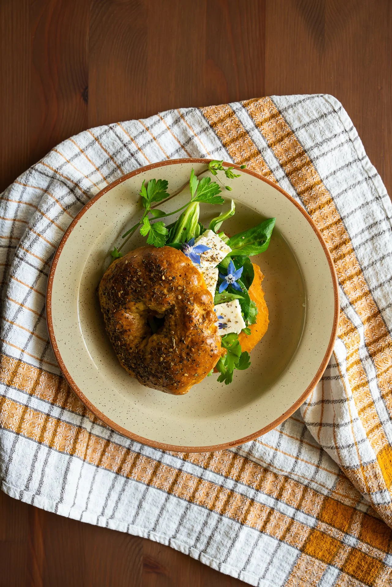 Bagel au zaatar, garni d'herbes fraîches, de feta, fleurs de bourrache, pickles d'oignons lactofermentés et tartinable de patate douce, le tout dans une assiette beige avec une bordure brune, placée sur un torchon blanc et orange. Vue du dessus.