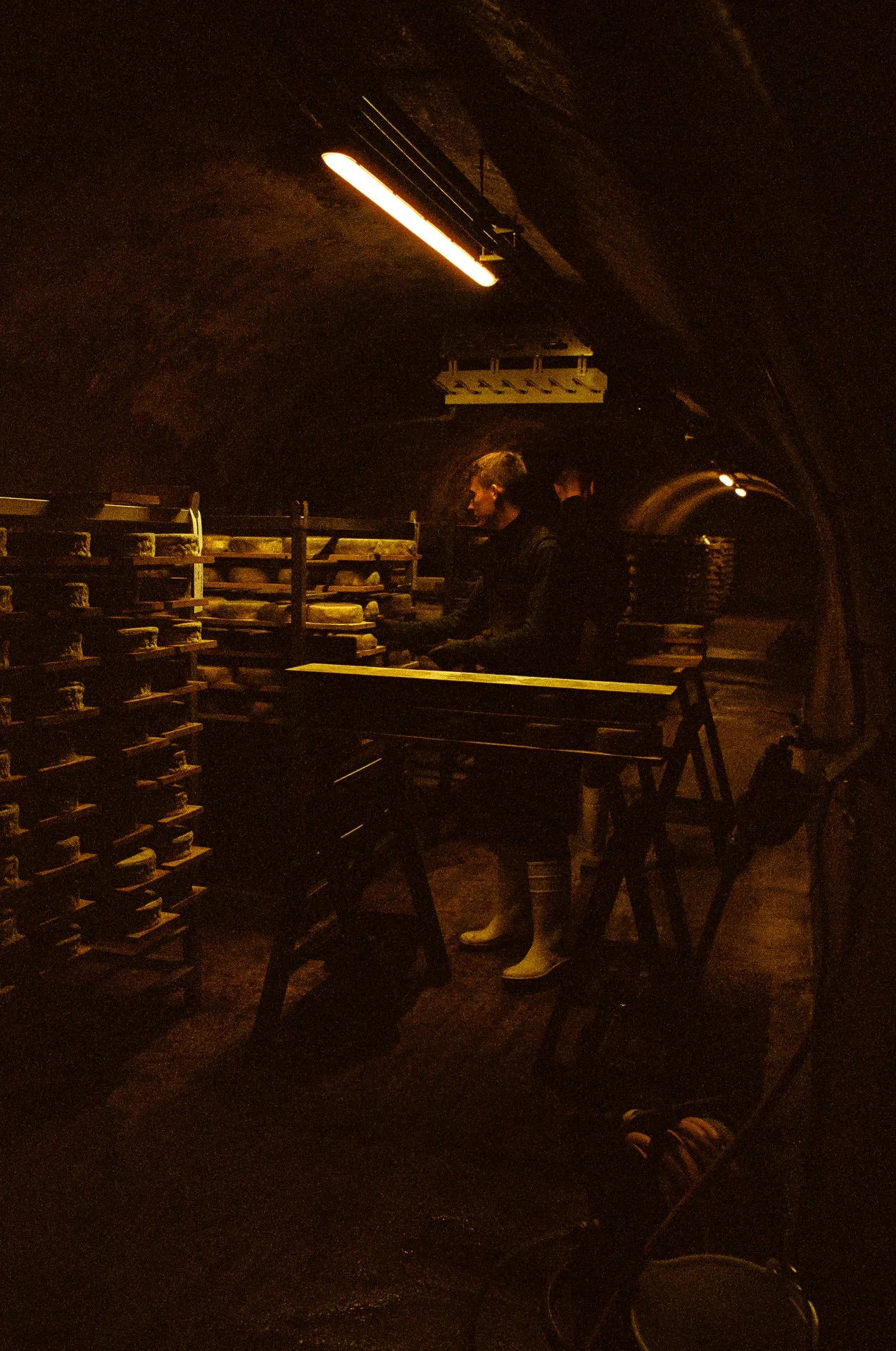 Une personne qui travaille dans une cave d'affinage de fromages, portant des bottes en caoutchouc, entourée de rangées de fromages en train d'affiner. Photographie argentique.