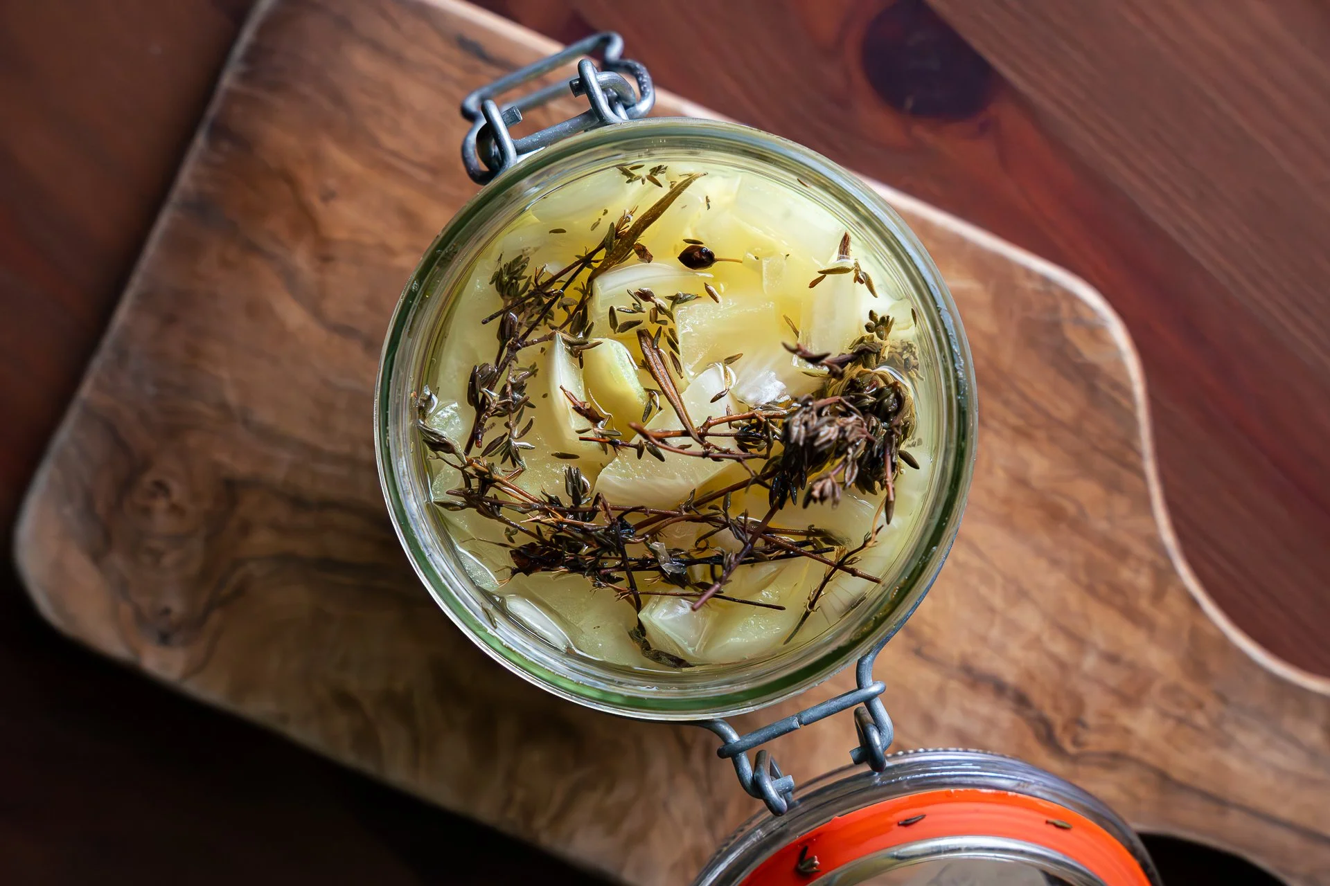 Pickles d'oignon jaune en bocal, avec branches de thym et romarin, vue du dessus, posé sur une planche en bois.