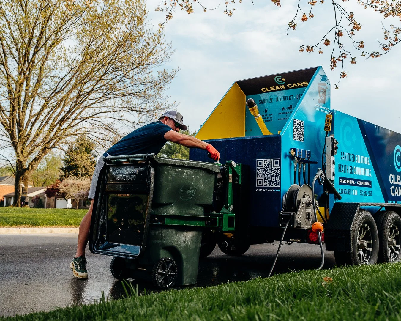 A man wearing a cap, sunglasses, and red gloves is cleaning a trash can with his truck on a suburban street with trees and houses in the background.