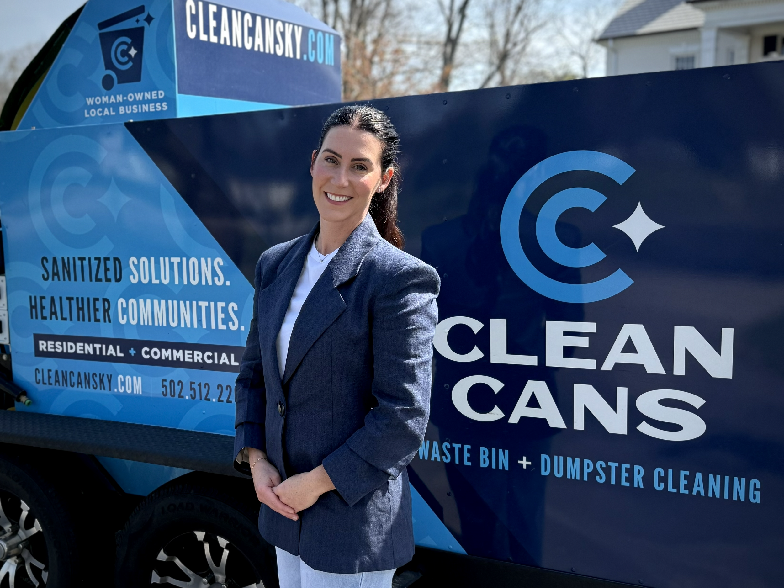 A woman, the owner in a gray blazer standing in front of a blue recycling truck with branding for cleaning services. She is smiling and has dark hair pulled back.