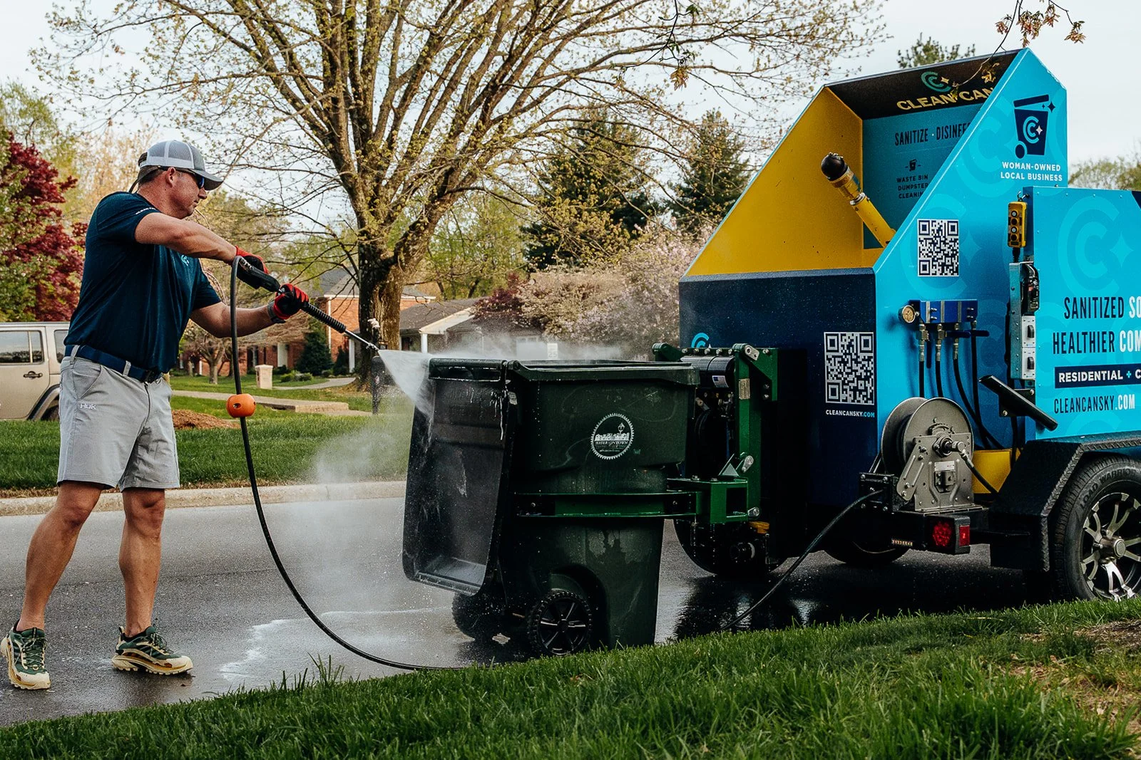 A man wearing a navy shirt, gray shorts, a gray cap, sunglasses, and gloves, is pressure washing his driveway with a portable trash can cleaning machine. The machine is blue and yellow, with QR codes and text promoting sanitization and local business