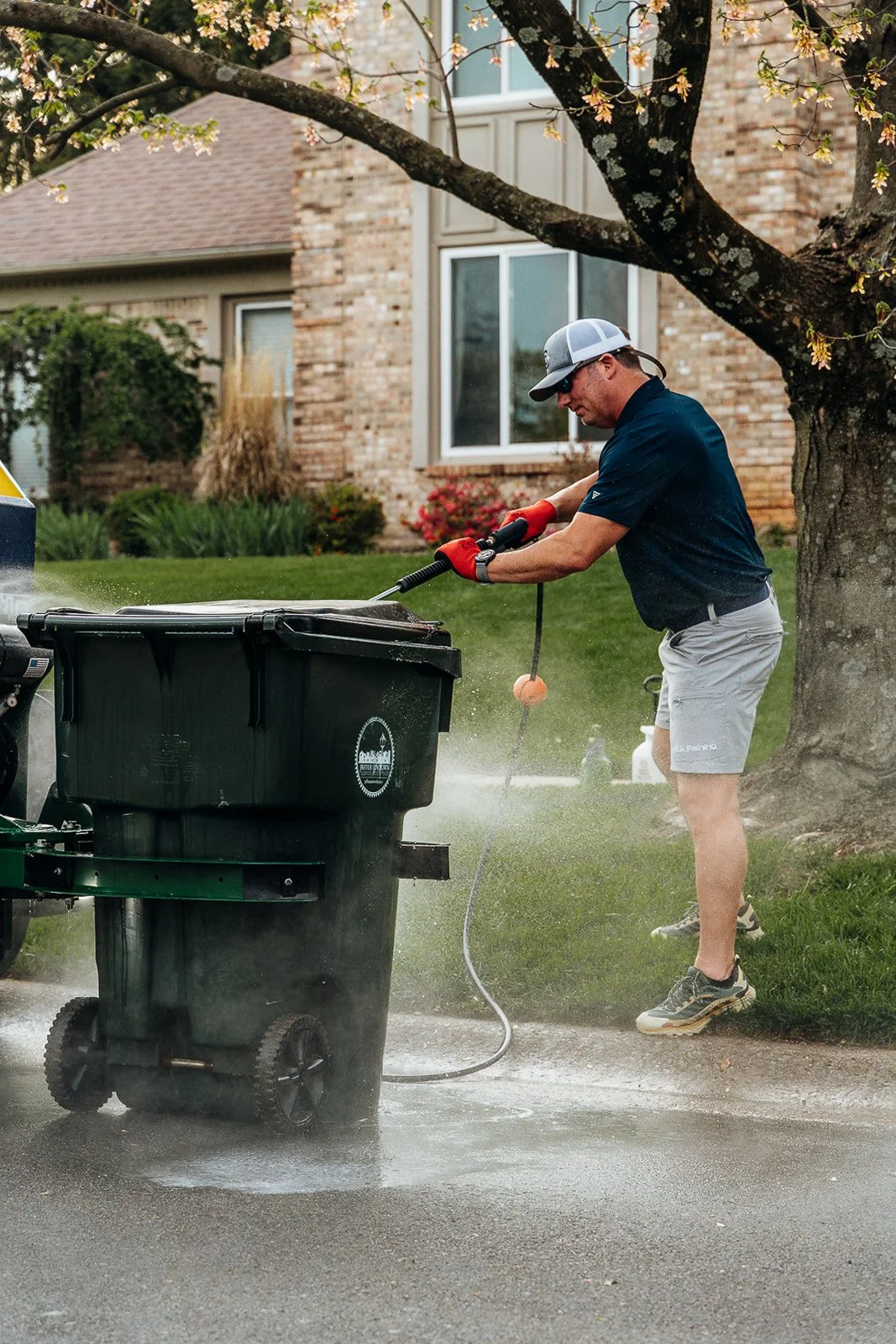 A man wearing a black shirt, gray shorts, and a cap is pressure washing a trash can with a hose, under a tree in a suburban neighborhood.