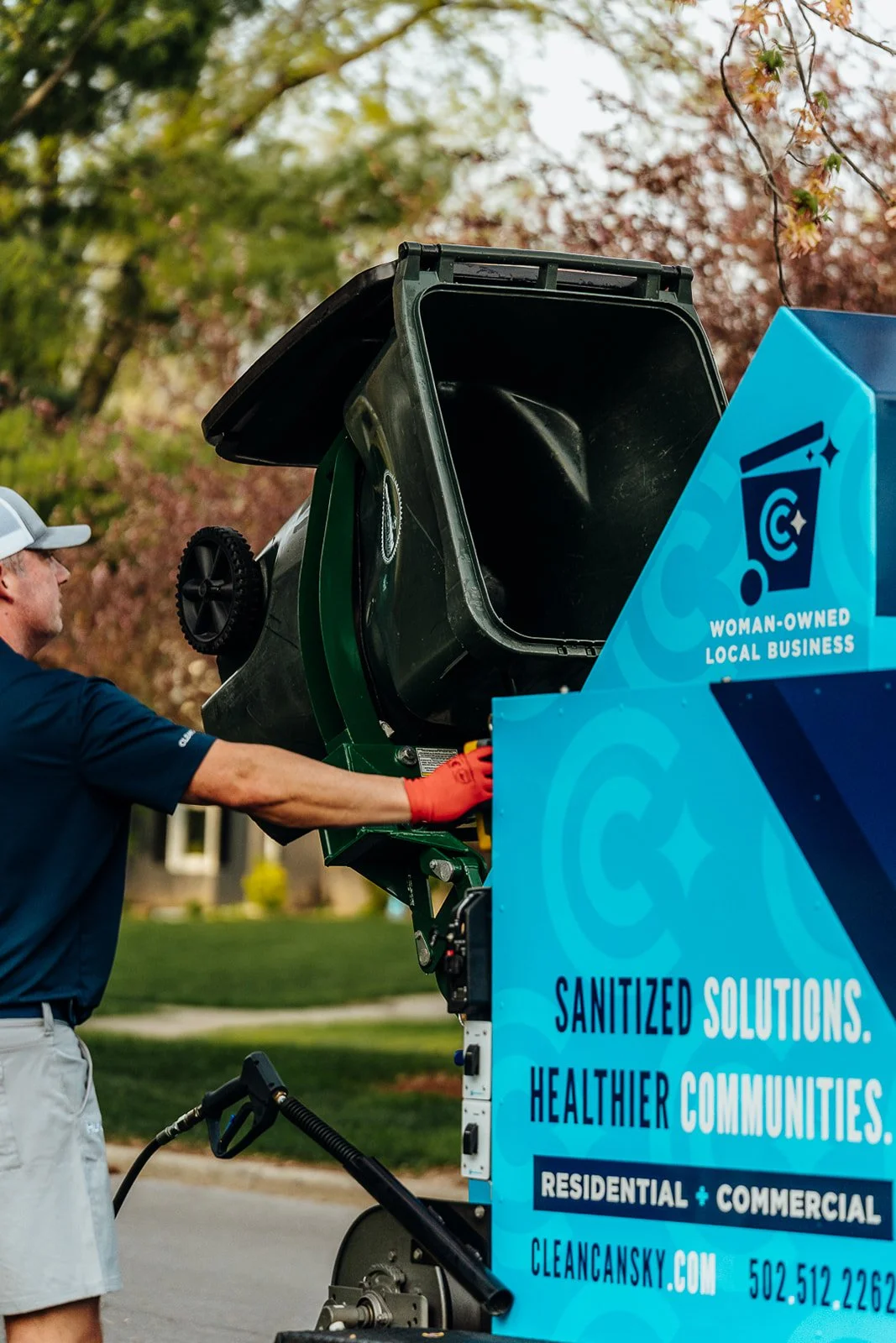A person in a blue shirt and white cap operating a trash can cleaning machine with a blue advertisement panel that reads "Sanitized solutions. Healthier communities."