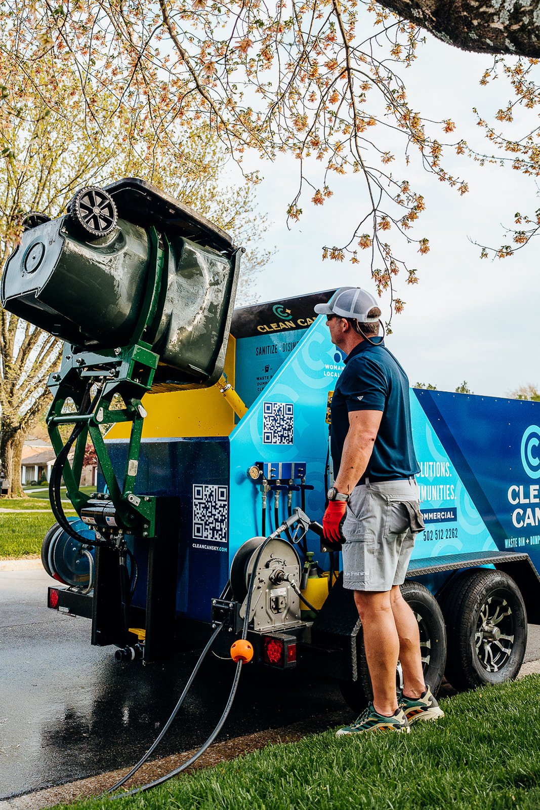 A man in casual attire, including a cap, sunglasses, and red gloves, stands next to a large mobile trash cleaning machine on a suburban street with trees and houses in the background.