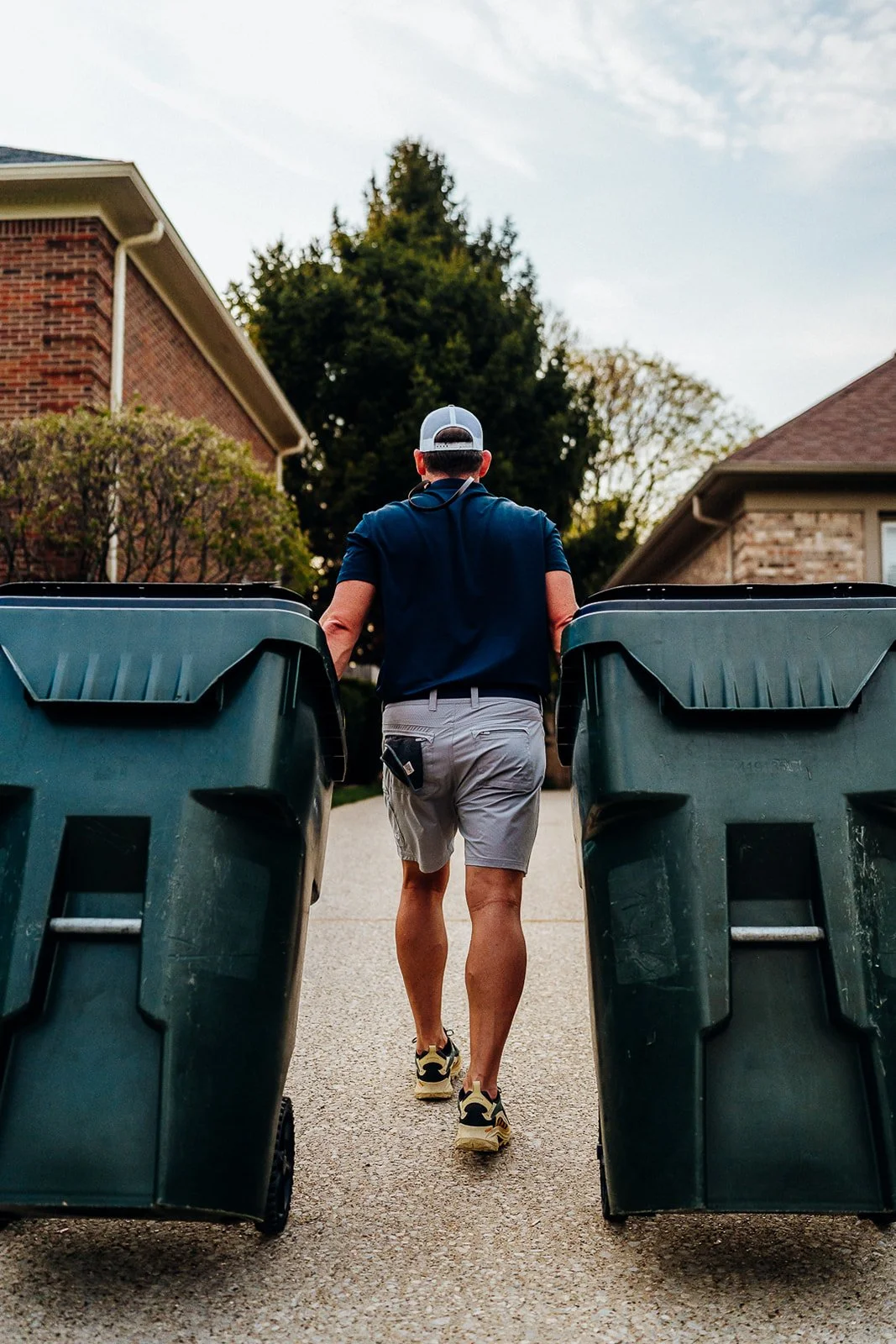 A man walking down an outdoor driveway between two large trash bins, with houses and trees visible in the background, during daytime.