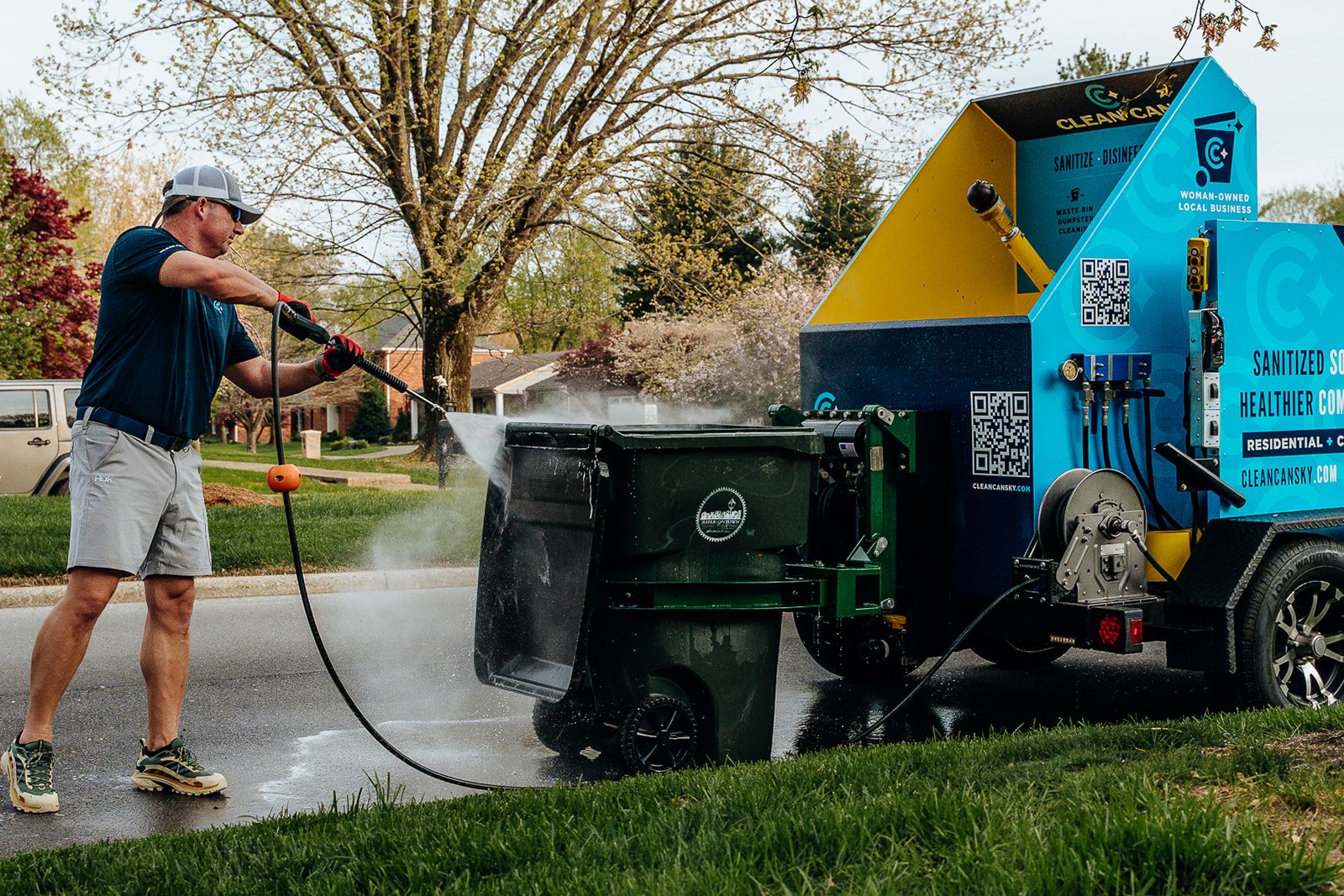 A man wearing a baseball cap, sunglasses, navy t-shirt, and gray shorts pressure washing a residential trash can with a large blue and yellow trash can cleaning machine.