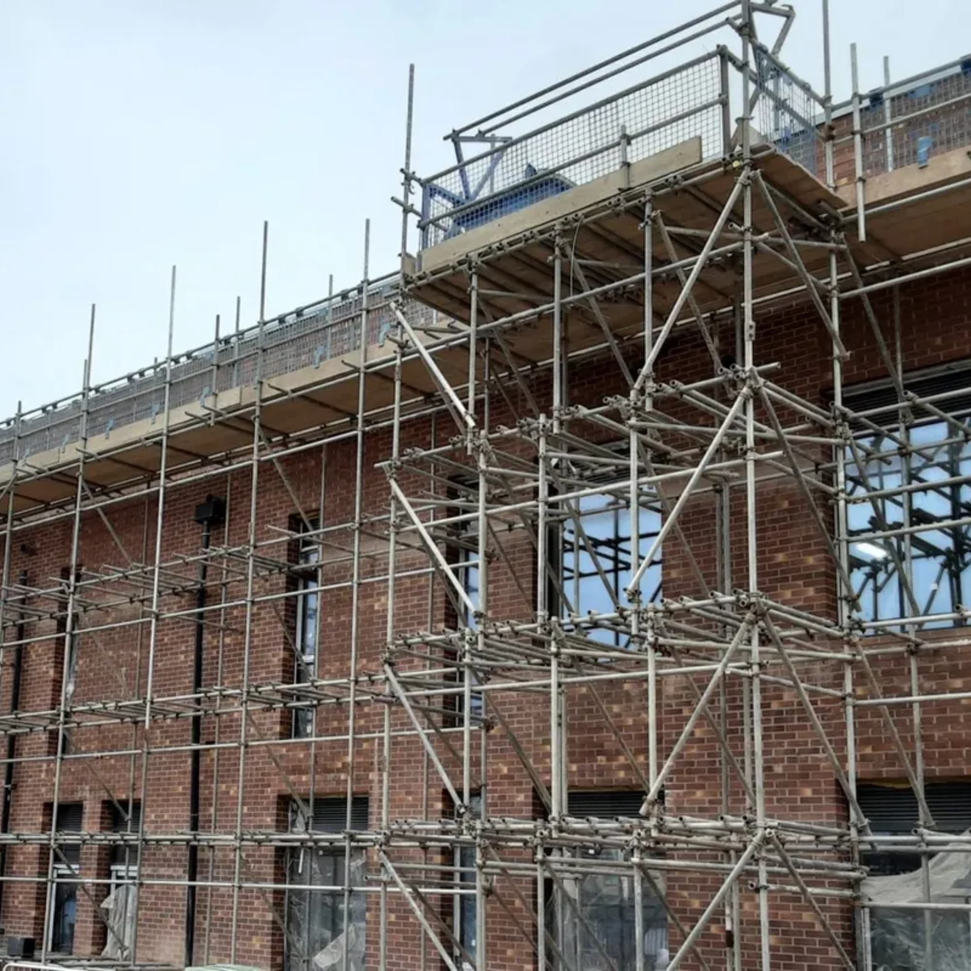 Construction scaffolding set up around a brick building with multiple windows, some covered with protective sheeting.