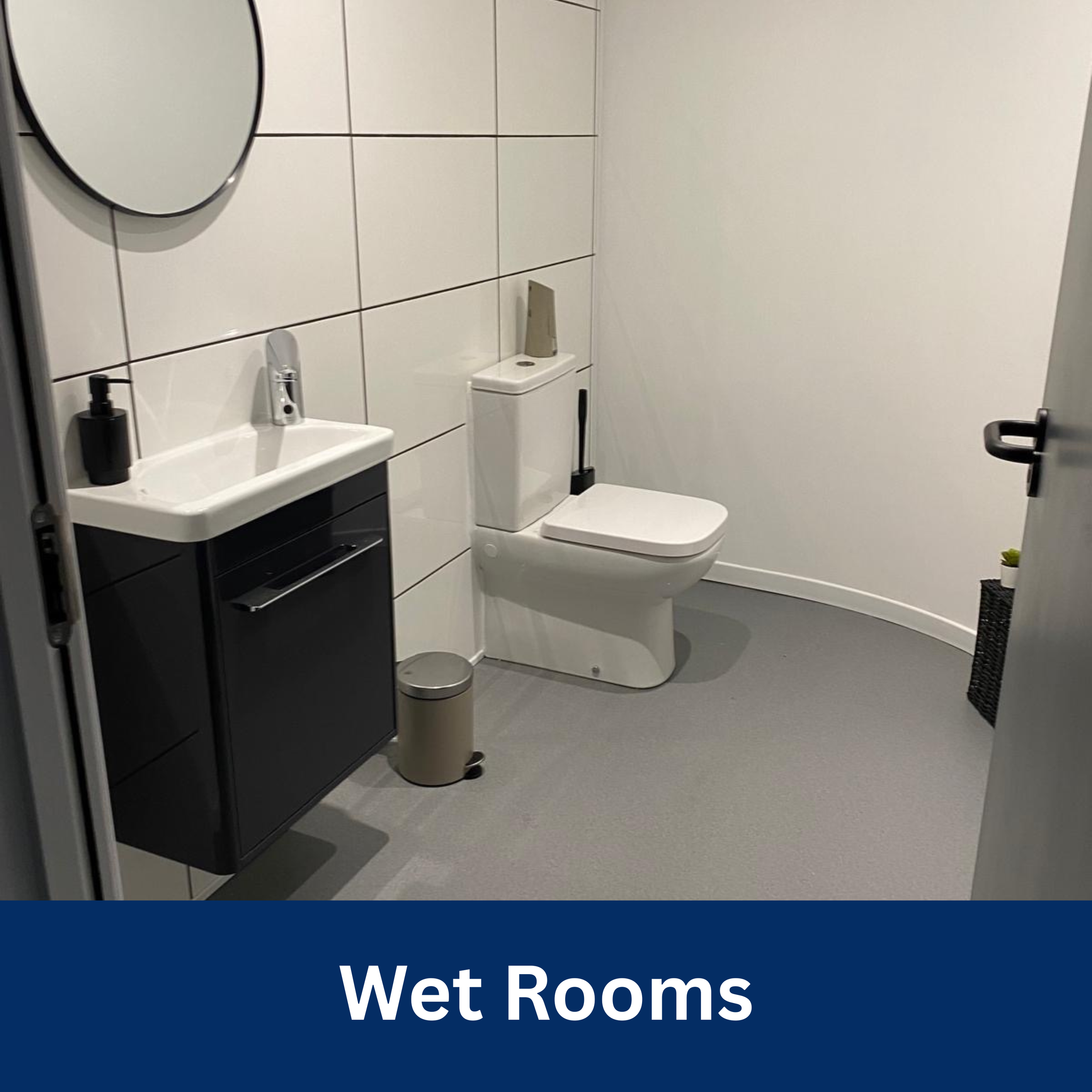 A white bathroom with a toilet, a black cabinet with a small sink, a round mirror, a black soap dispenser, and a small trash can. The walls are tiled in white, and the floor is gray. There is a plant in a black basket in the corner.