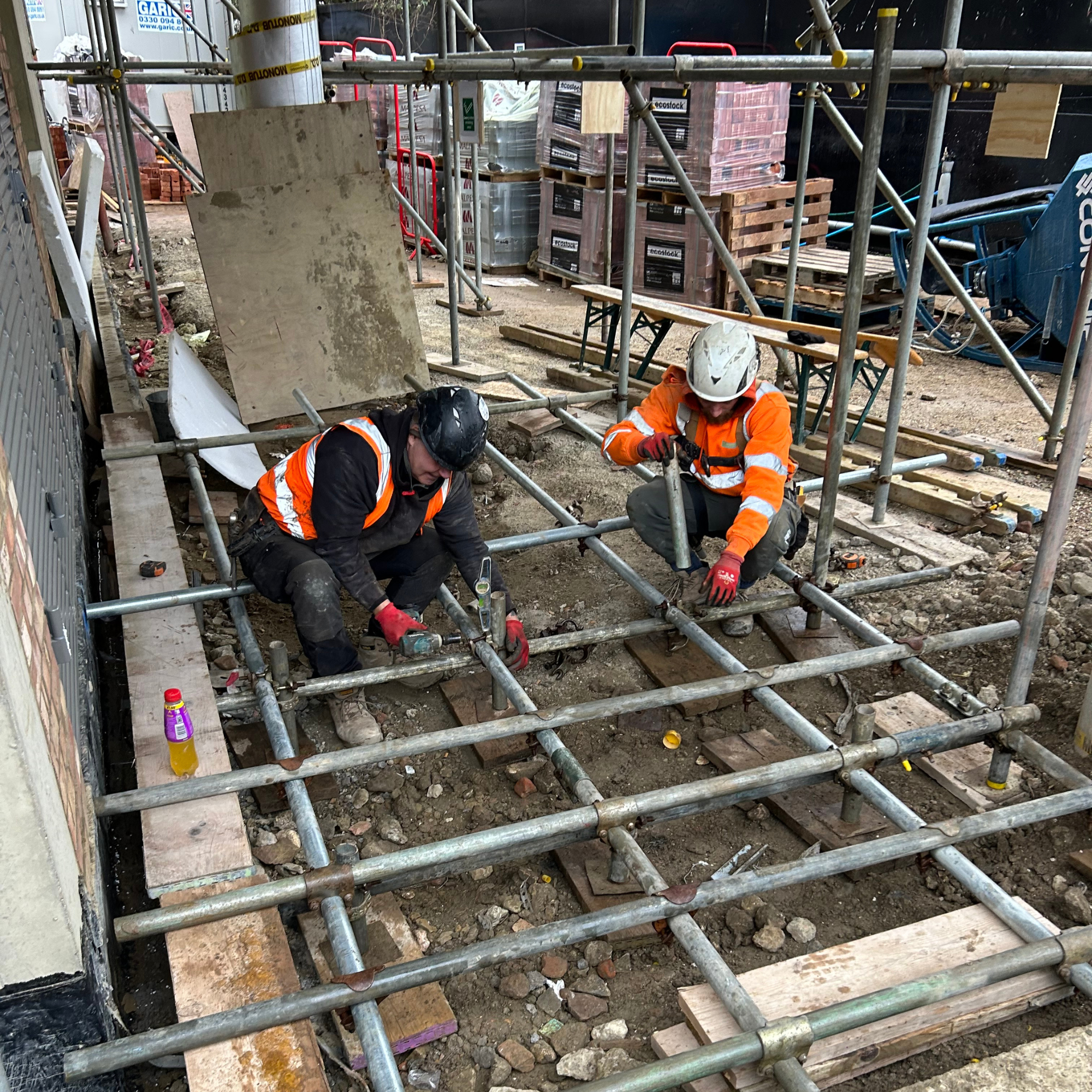 Two construction workers wearing helmets and orange safety vests working on a scaffolding structure at a construction site. They are handling tools and working on the ground, surrounded by construction materials and equipment.