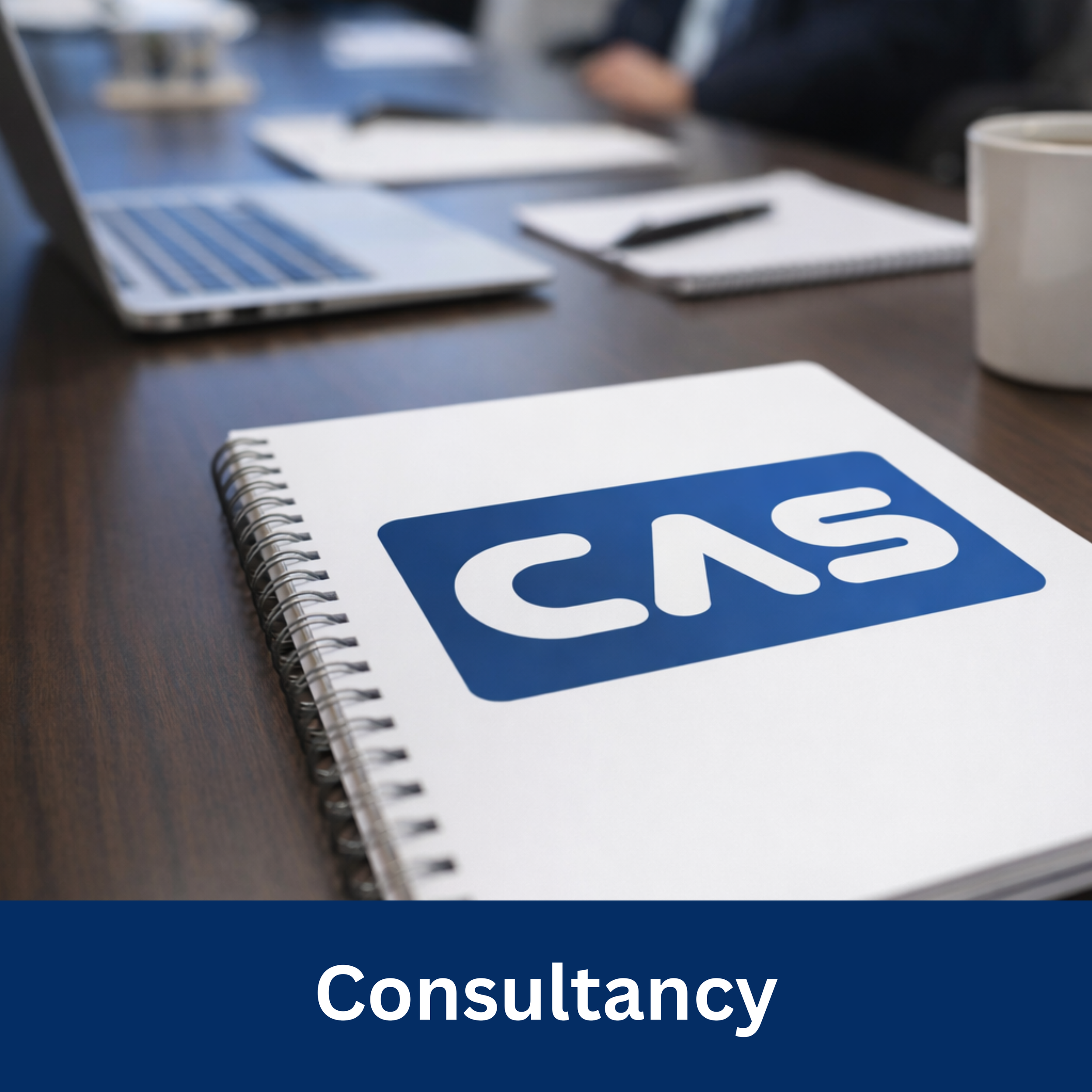 Notebook with 'CAS' logo on cover on a wooden table with laptops, notebooks, and a coffee mug in the background.
