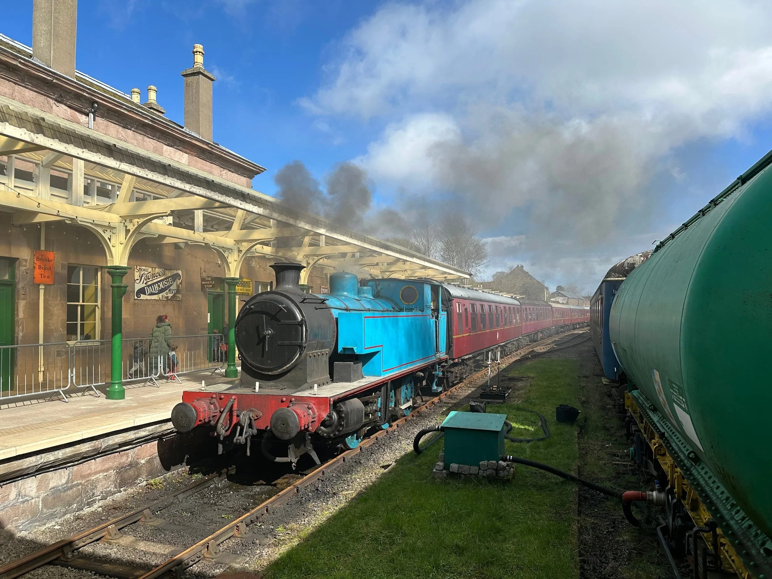 A vintage train station with a blue steam locomotive, red train cars, and a water tank in a scenic outdoor setting.