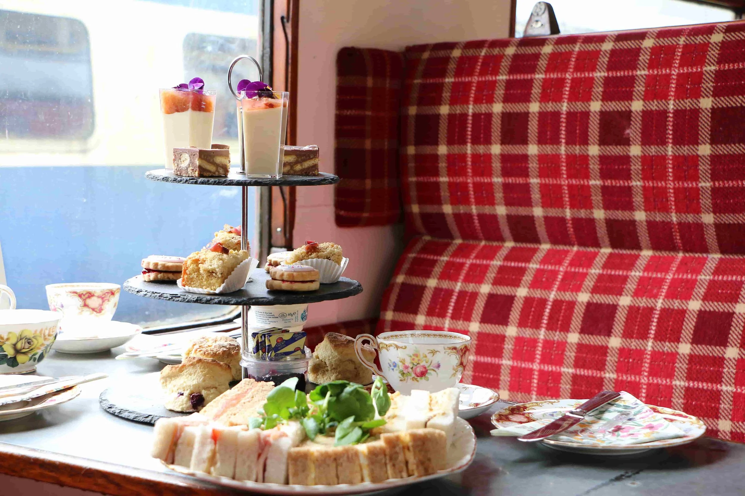 A table set with assorted desserts including cakes, cookies, and finger sandwiches, with a tiered tray holding more treats, in front of a red checkered booth at a cafe or restaurant.