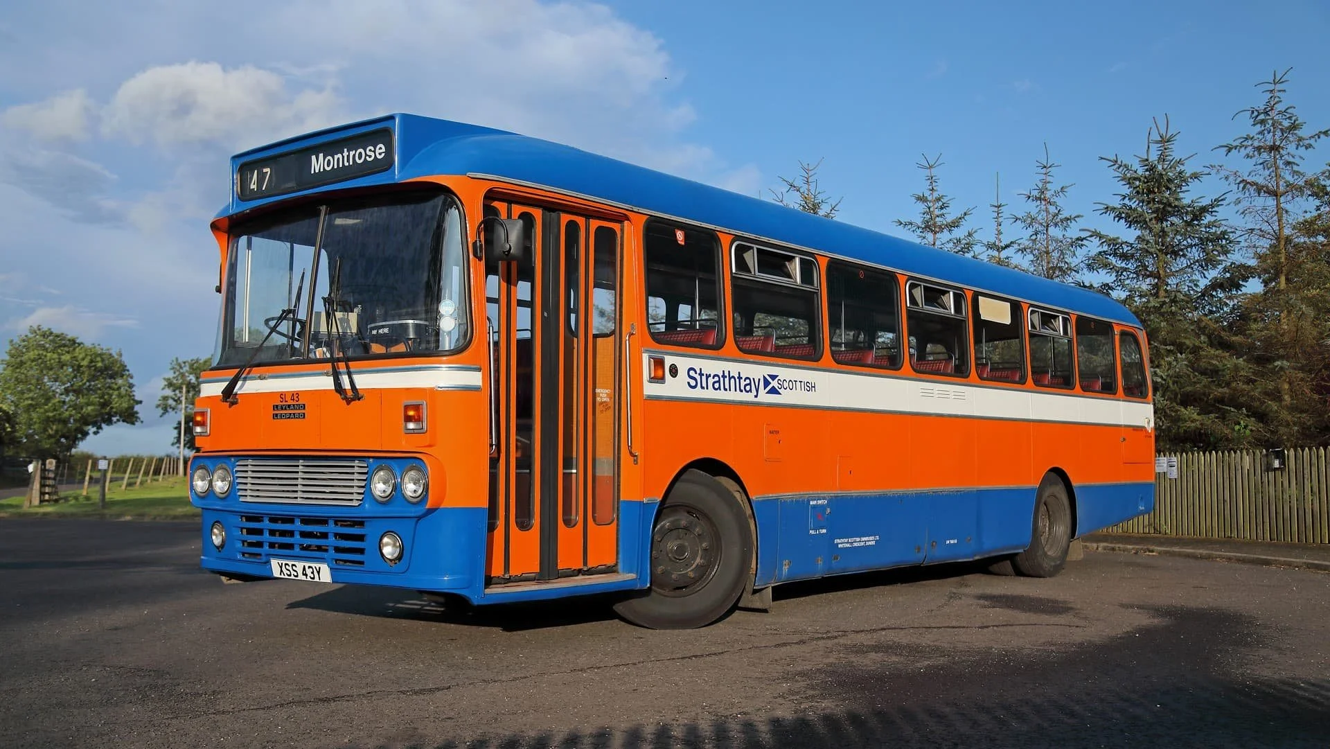 A vintage bus painted in orange, white, and blue colors with the route destination 'Montrose' displayed on the front. The bus has the logo and name 'Strathclyde Scottish' on its side, with several windows and a door near the front. The background features a partly cloudy sky, green trees, and a wooden fence.