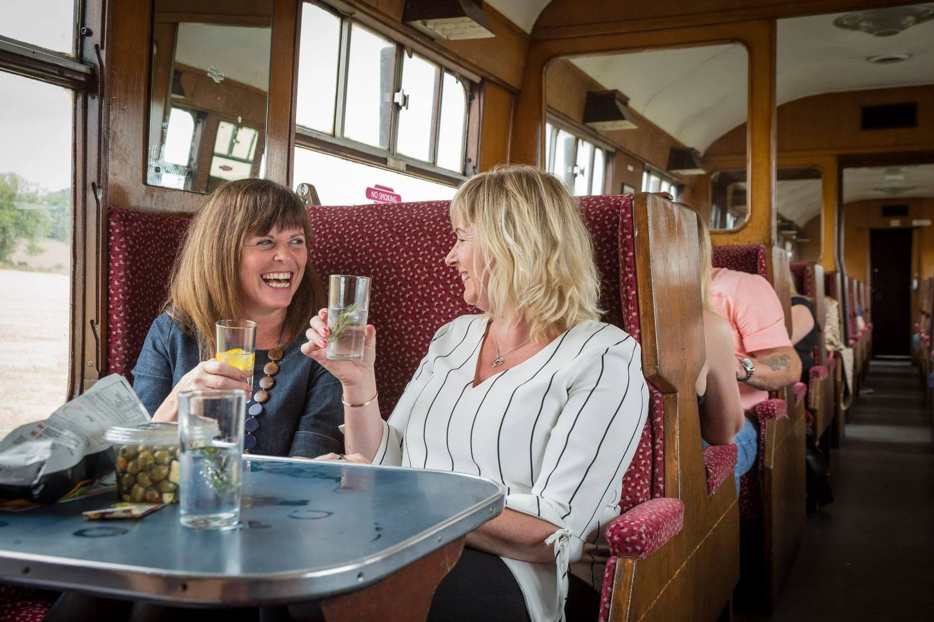 Women enjoying drinks and conversation on a train with wood-paneled walls and red-patterned seats.