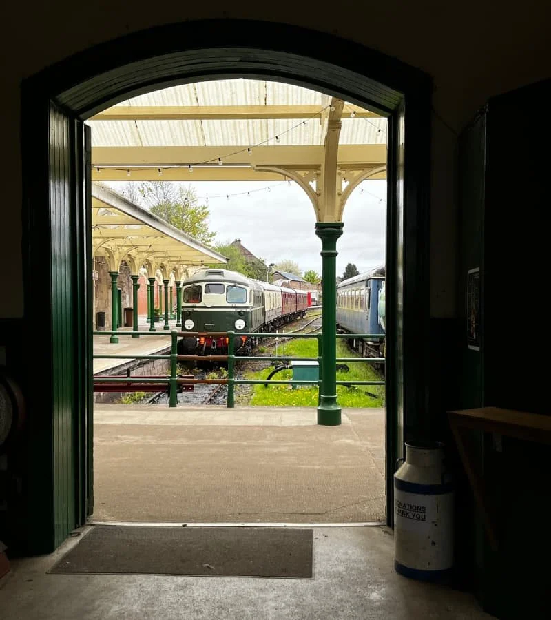 View of Caledonian Railway, Brechin train station through an arched doorway, with a vintage train on the tracks and a platform with decorative columns and string lights.