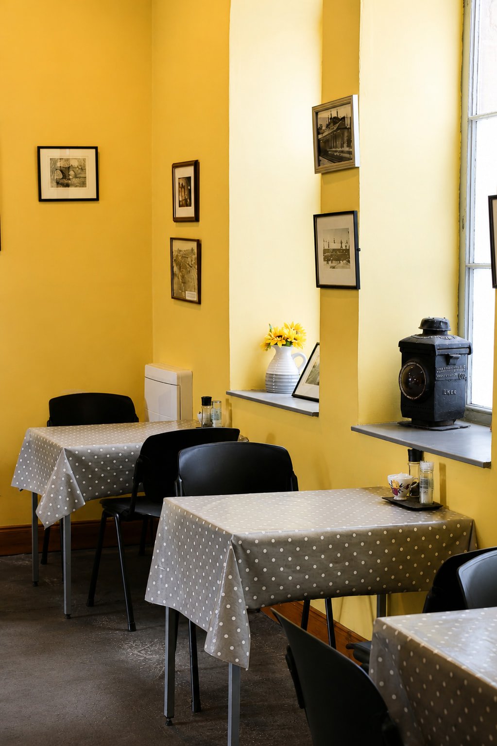 A cozy dining area with yellow walls, decorated with black and white framed pictures, three tables covered with gray polka dot tablecloths, black chairs, sunflower in a white pitcher, a vintage camera, and sunlight streaming through a window.