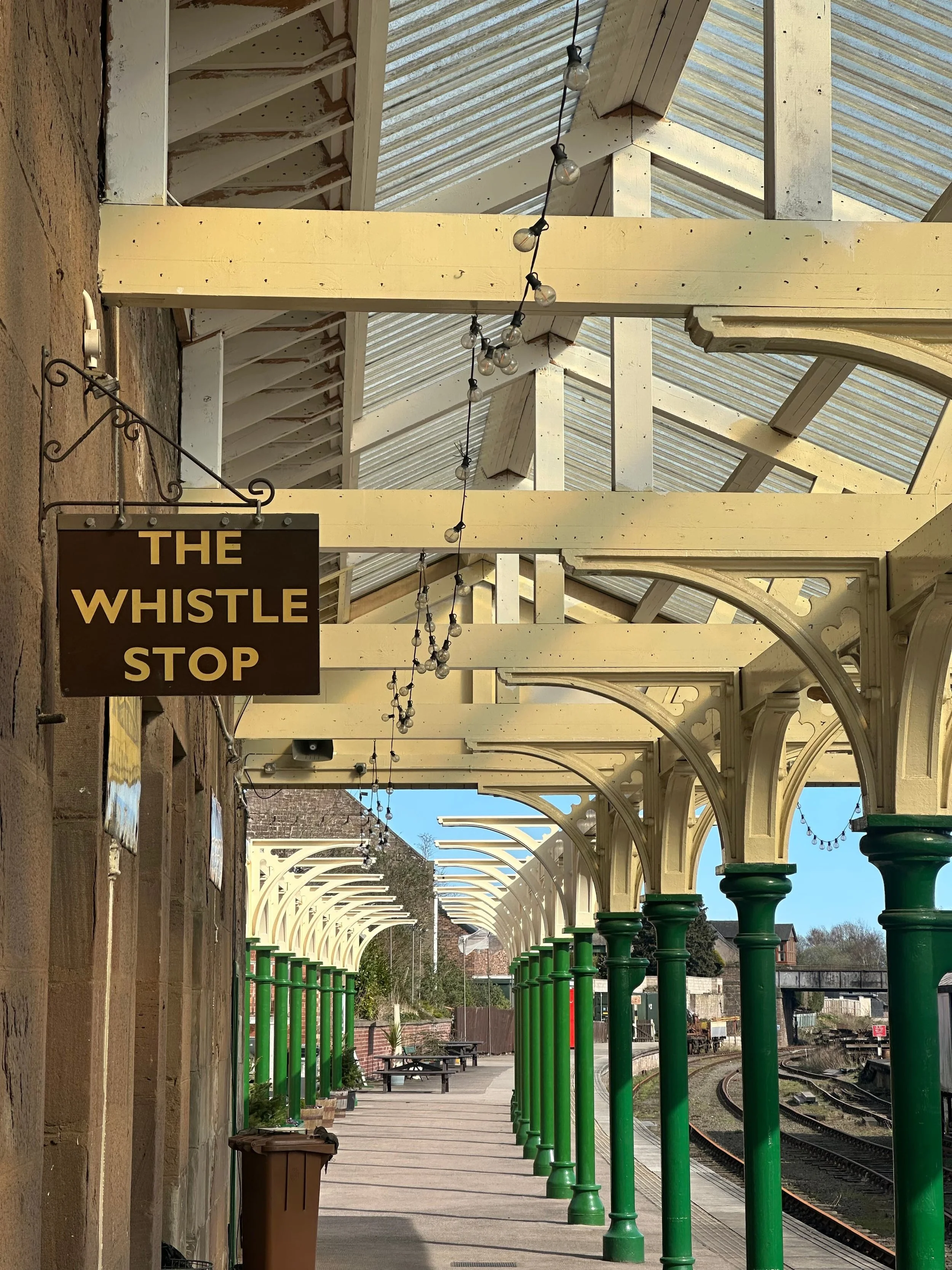 View of Caledonian Railway, Brechin Station platform with green support columns and an arched roof structure. A sign reads 'The Whistle Stop' for the cafe. A string of light bulbs hangs above the platform. The tracks are visible on the right side.