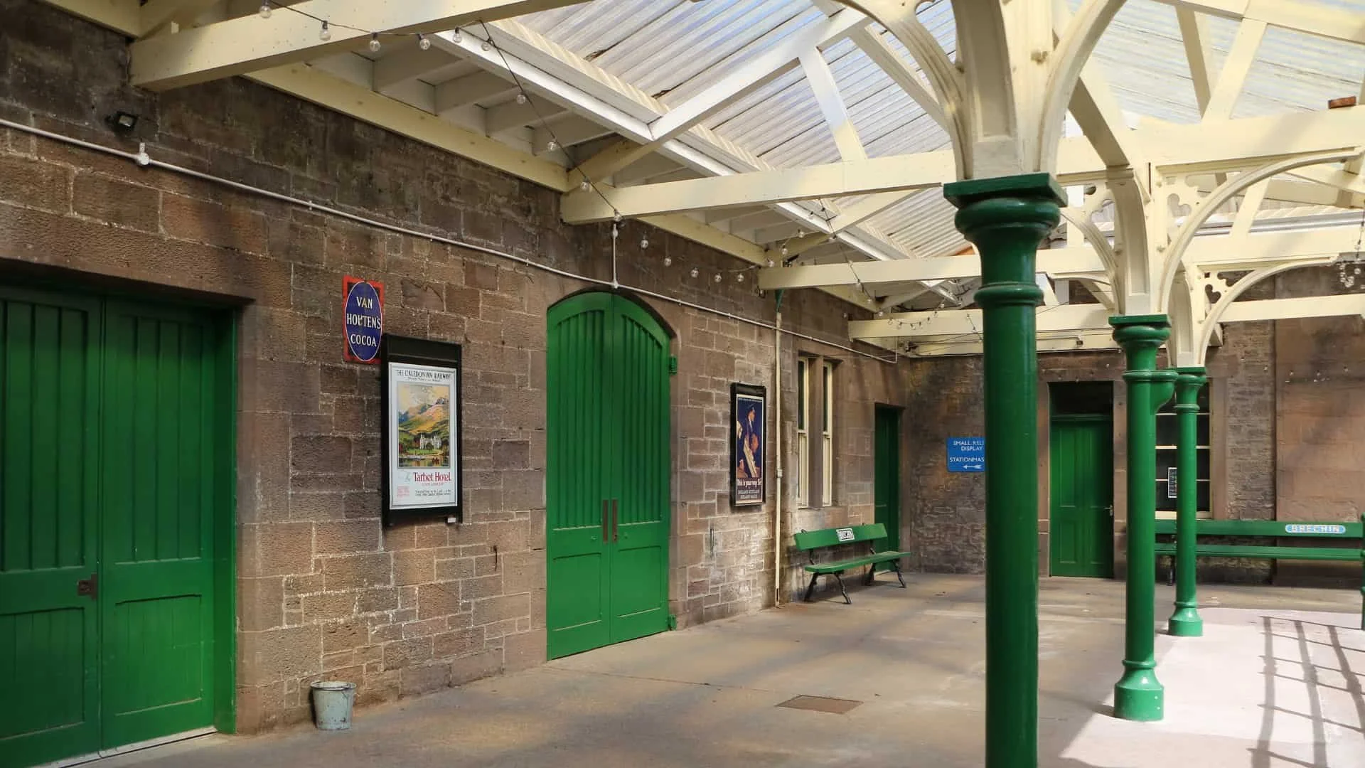 A vintage train station platform with green doors, informational posters, a bench, and green metal columns supporting a skylight roof, with string lights hanging overhead.