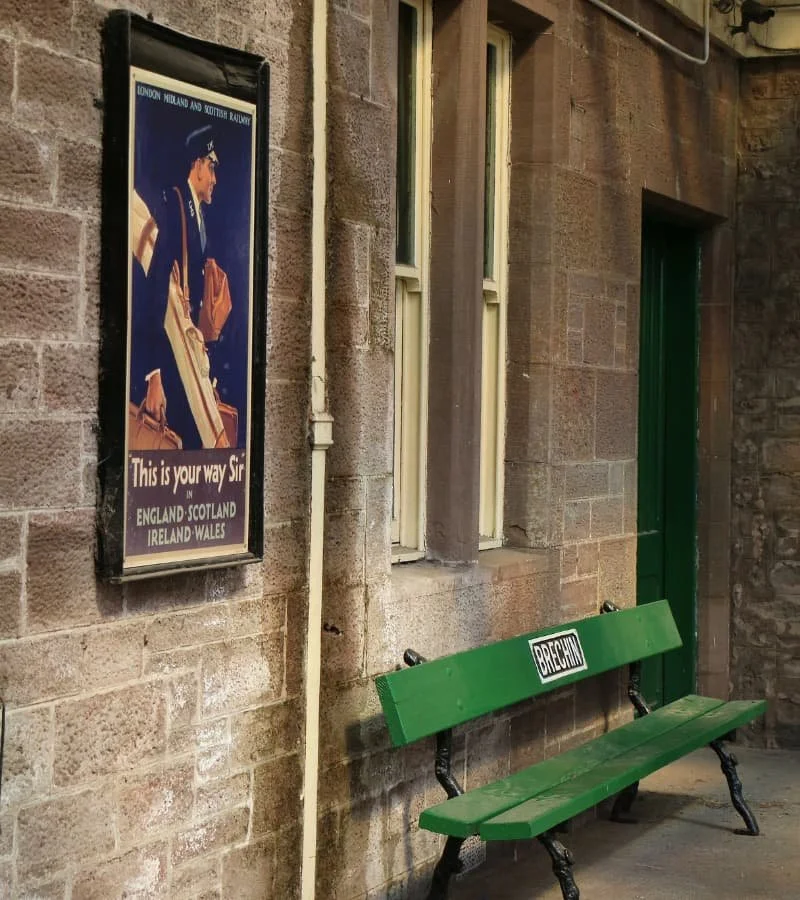 A vintage train station poster on a brick wall shows a conductor in uniform, with text indicating a route from England, Scotland, Ireland, and Wales. Below the poster is a green bench with a sign reading "Brechin," situated against the wall near a window and a green door.