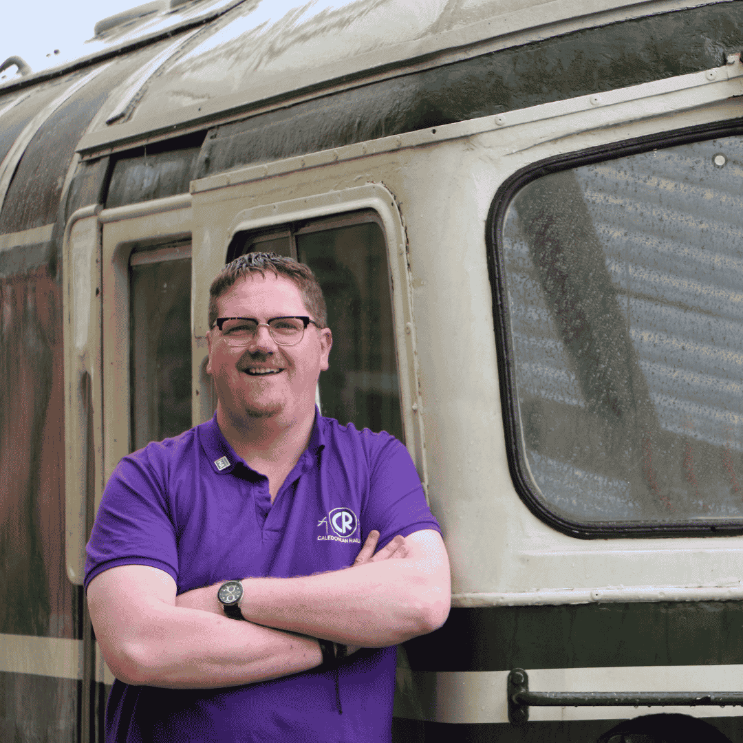 A man smiling with glasses and a purple shirt standing next to an old, weathered train carriage.