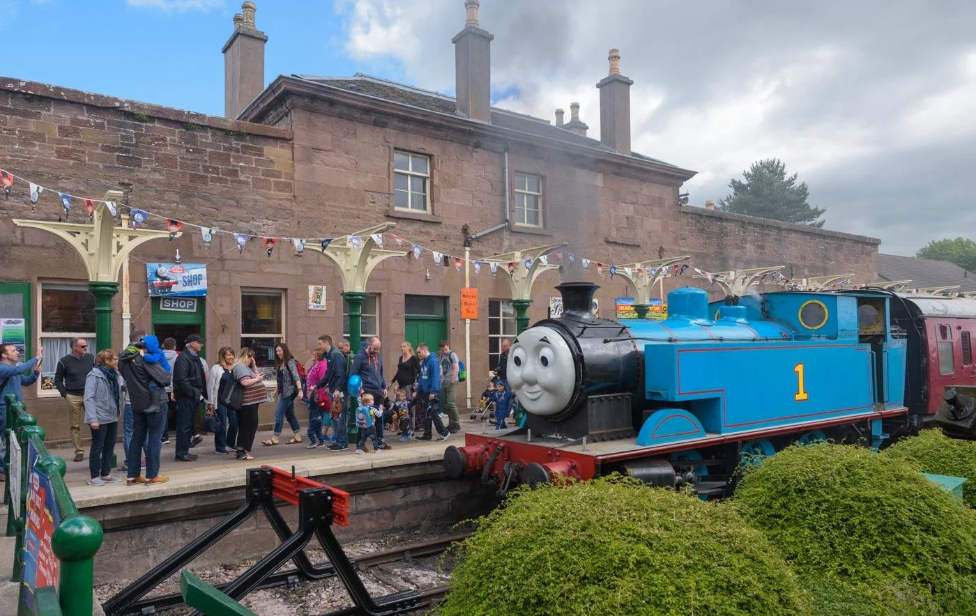 A crowd of people walking along a platform at Caledonian Railway, Brechin train station with a large animated blue train resembling Thomas the Tank Engine on the tracks nearby.