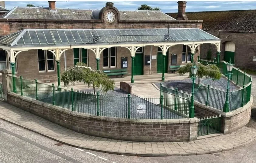 Small historic train station with a glass awning and a clock on the roof, surrounded by a low brick wall and green fencing, with benches and trees outside.