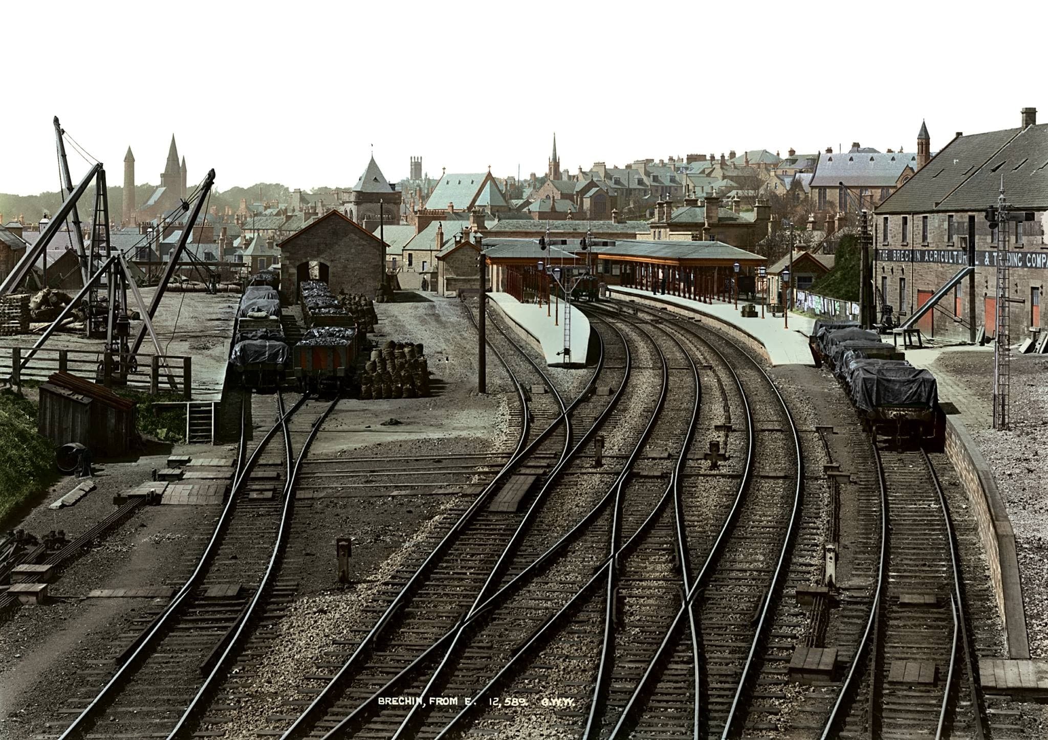 View of Caledonian Railway station with multiple train tracks and a station platform, surrounded by the town of Brechin, Angus with buildings and church steeples in the background.