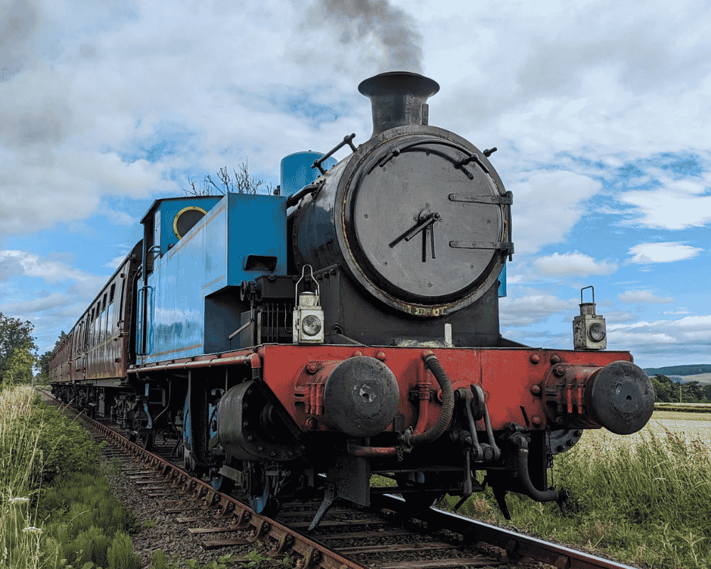 Thomas the Tank Engine train on Caledonian Railway railroad tracks with green grass and fields, cloudy sky in the background.