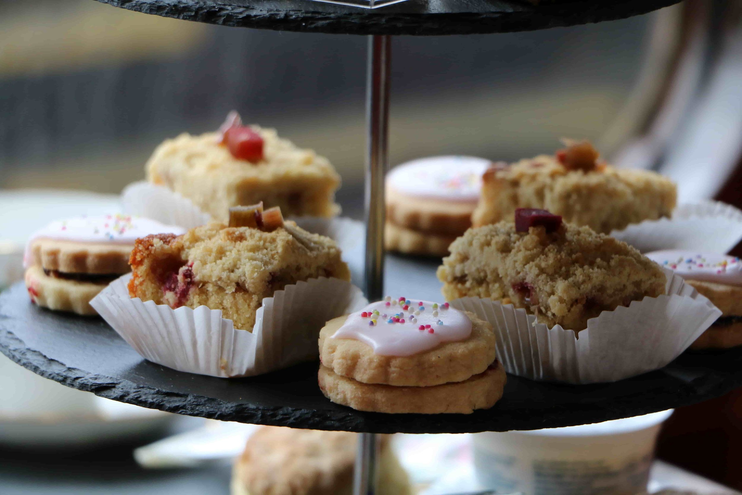 Assorted baked goods on a two-tiered black tray, including cookies and cake slices, some decorated with icing and colorful sprinkles.