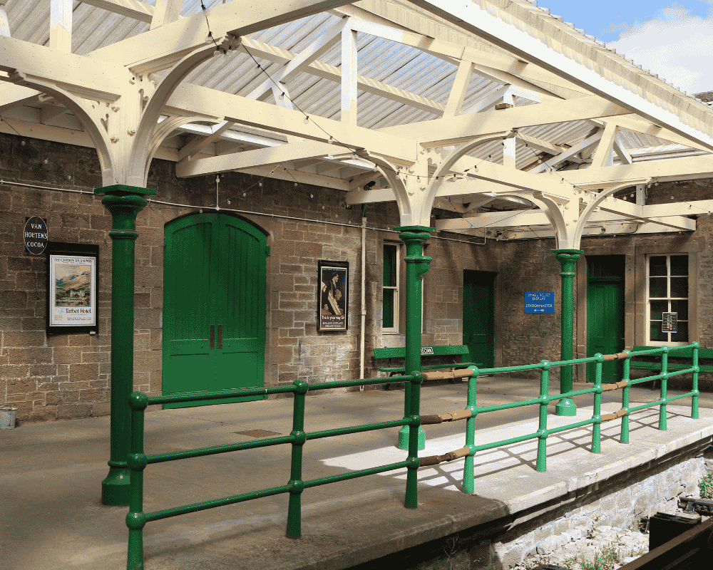 Old brick train station platform with green doors, benches, and a canopy supported by green columns.