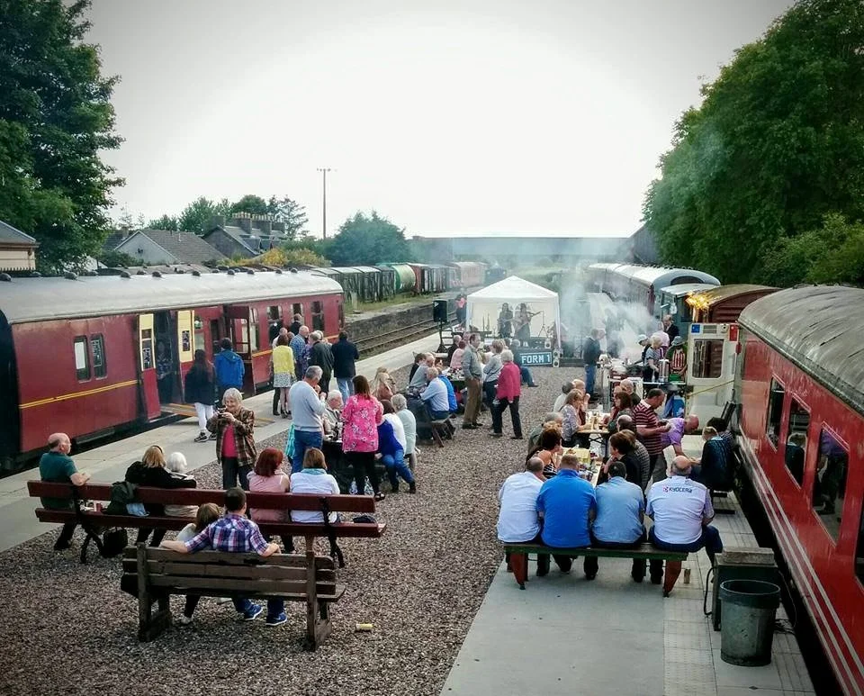 People gathered at a train station for an outdoor event with a stage, food, and drinks.