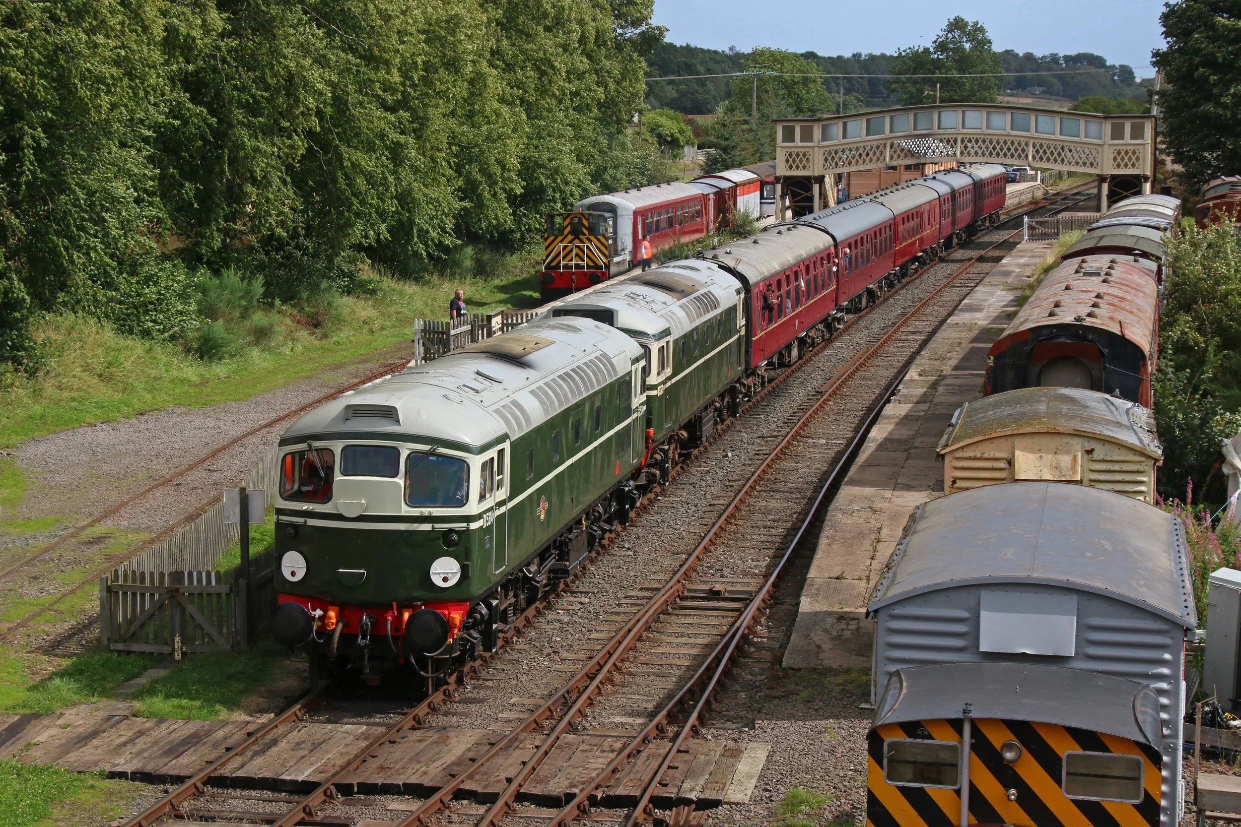 A passenger train with a green locomotive and red cars on a railway track, passing under a pedestrian bridge in a rural area with dense green trees on both sides.