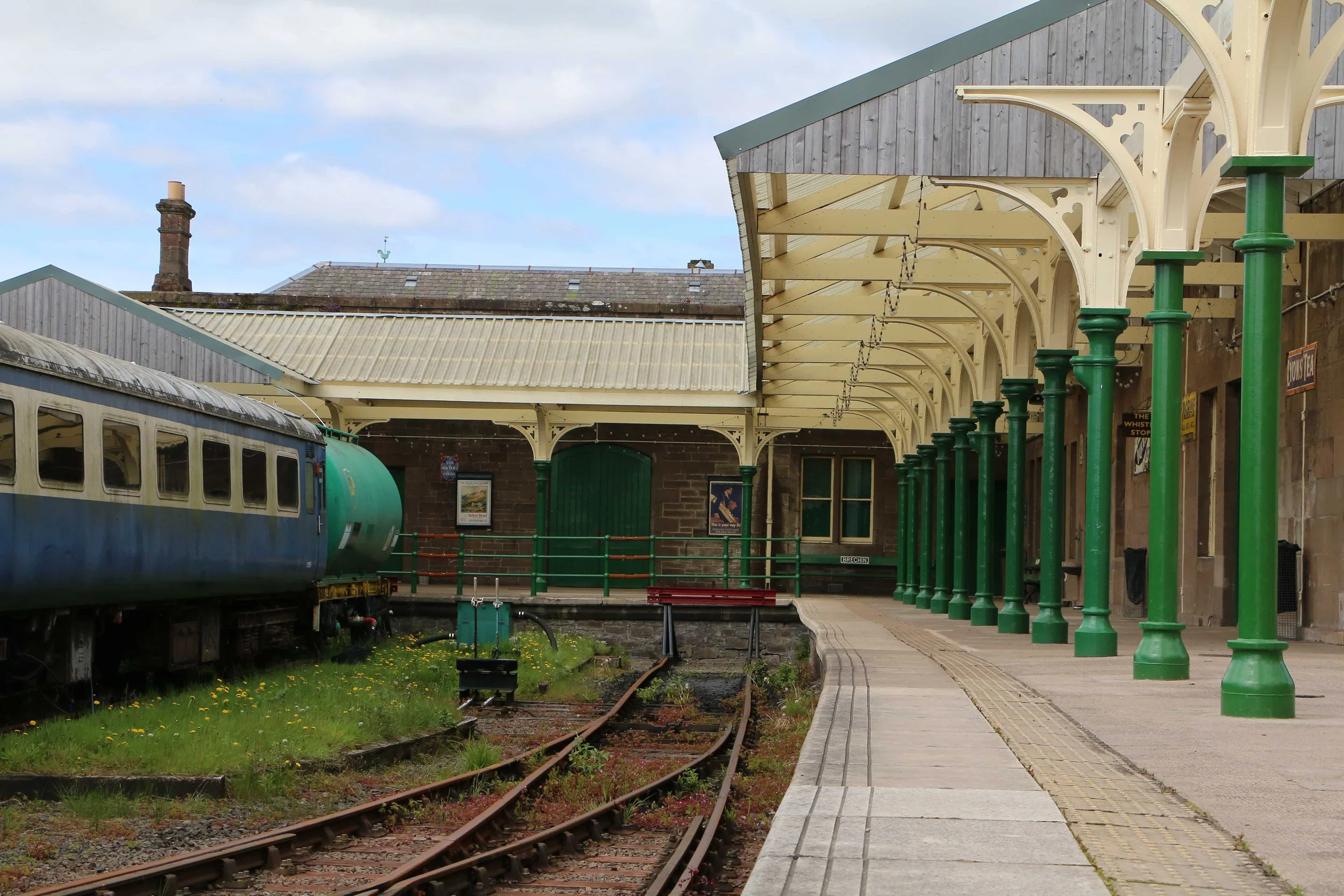 Caledonian Railway, Brechin station platform with a blue train on the left, a brick building with a covered waiting area on the right, and railway tracks running through the center. The platform has green support columns and decorative arches.