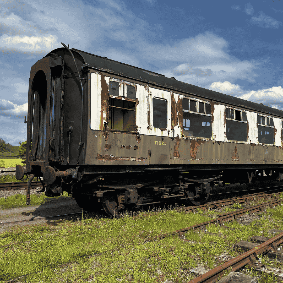 Old rusted train car on railway tracks in a grassy field under a partly cloudy sky.