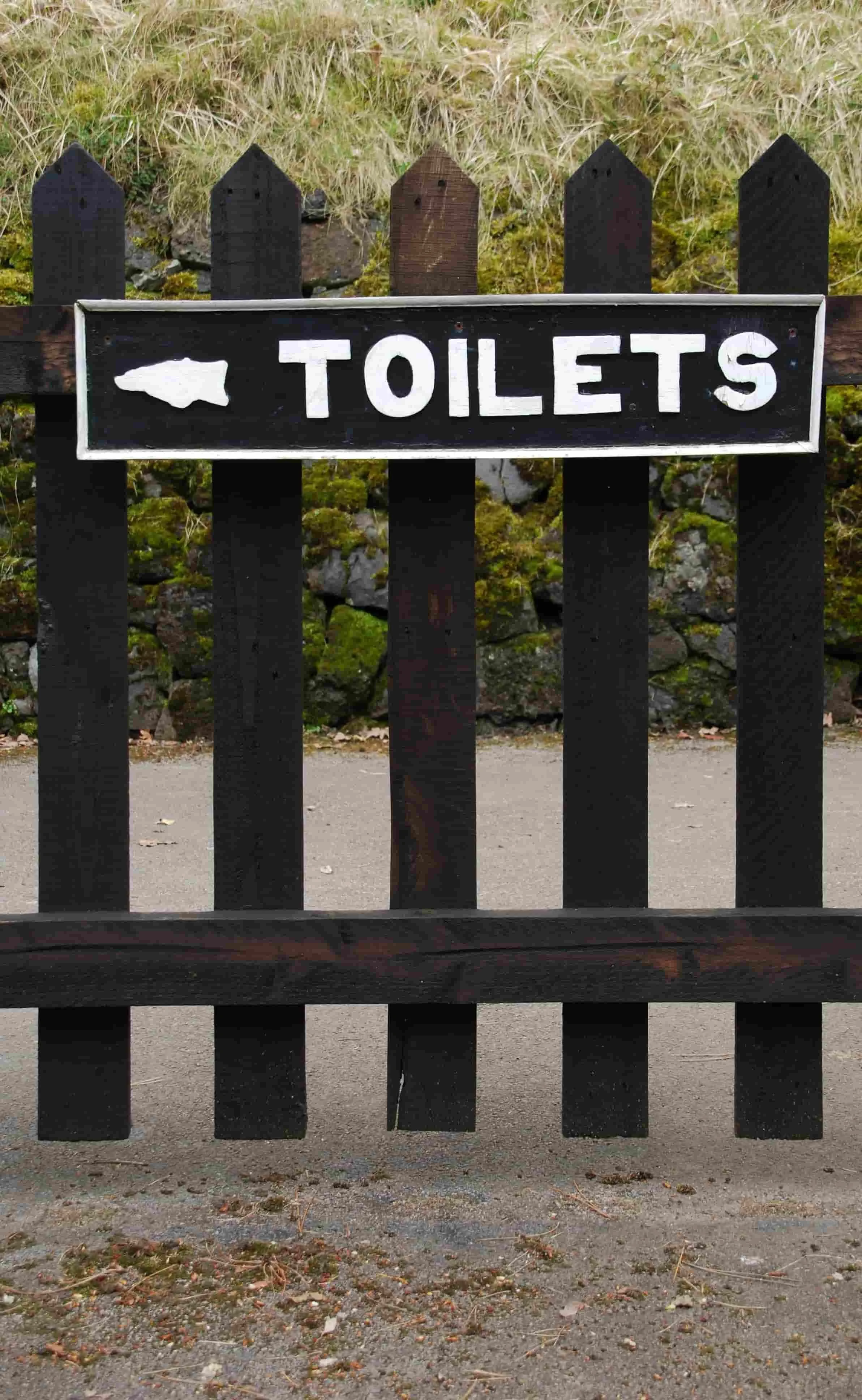 Wooden fence with a sign indicating the direction to toilets, outdoors on a gravel path with grass and moss in the background.