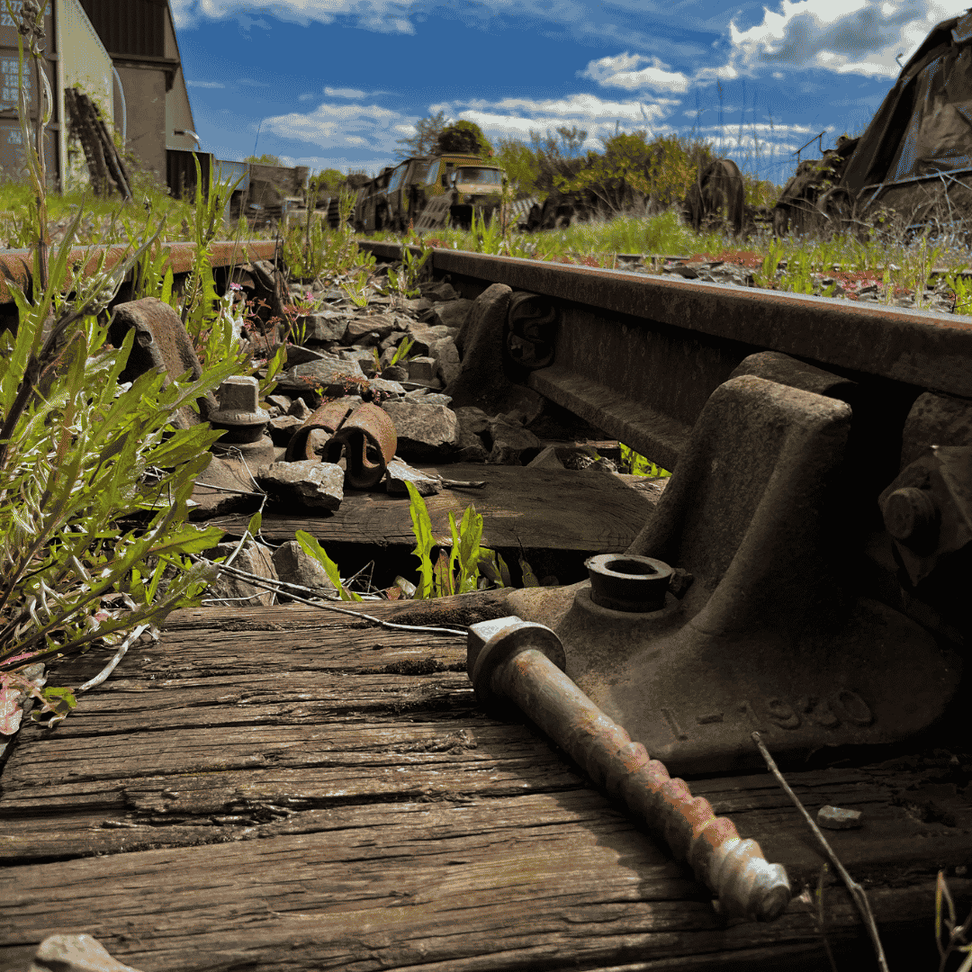 Close-up of an abandoned train track with rusted rails and overgrown vegetation, with old train cars and a partly cloudy sky in the background.