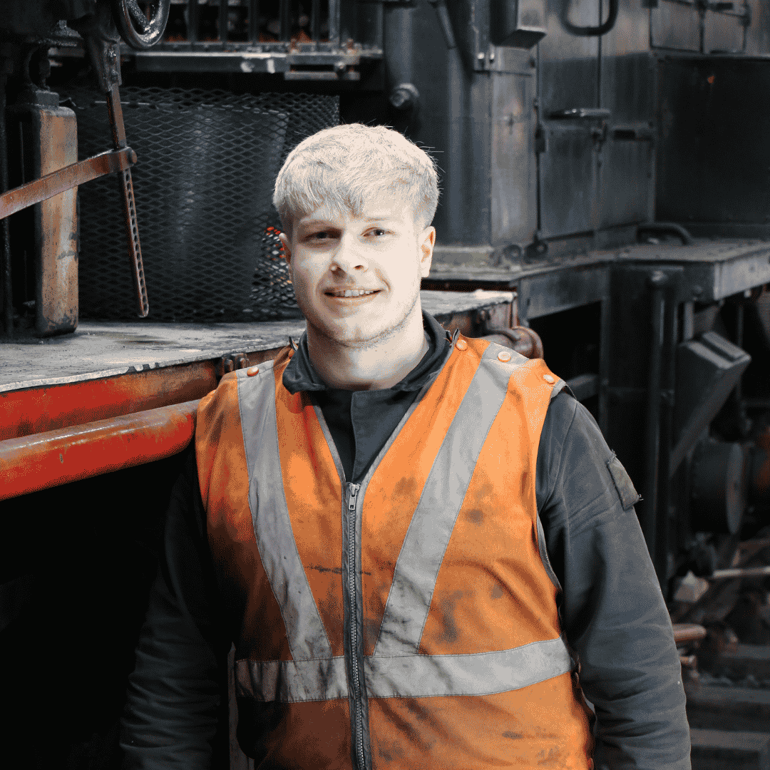 Young man with blond hair, wearing a black jacket and orange reflective safety vest, standing in front of a vintage black train engine.