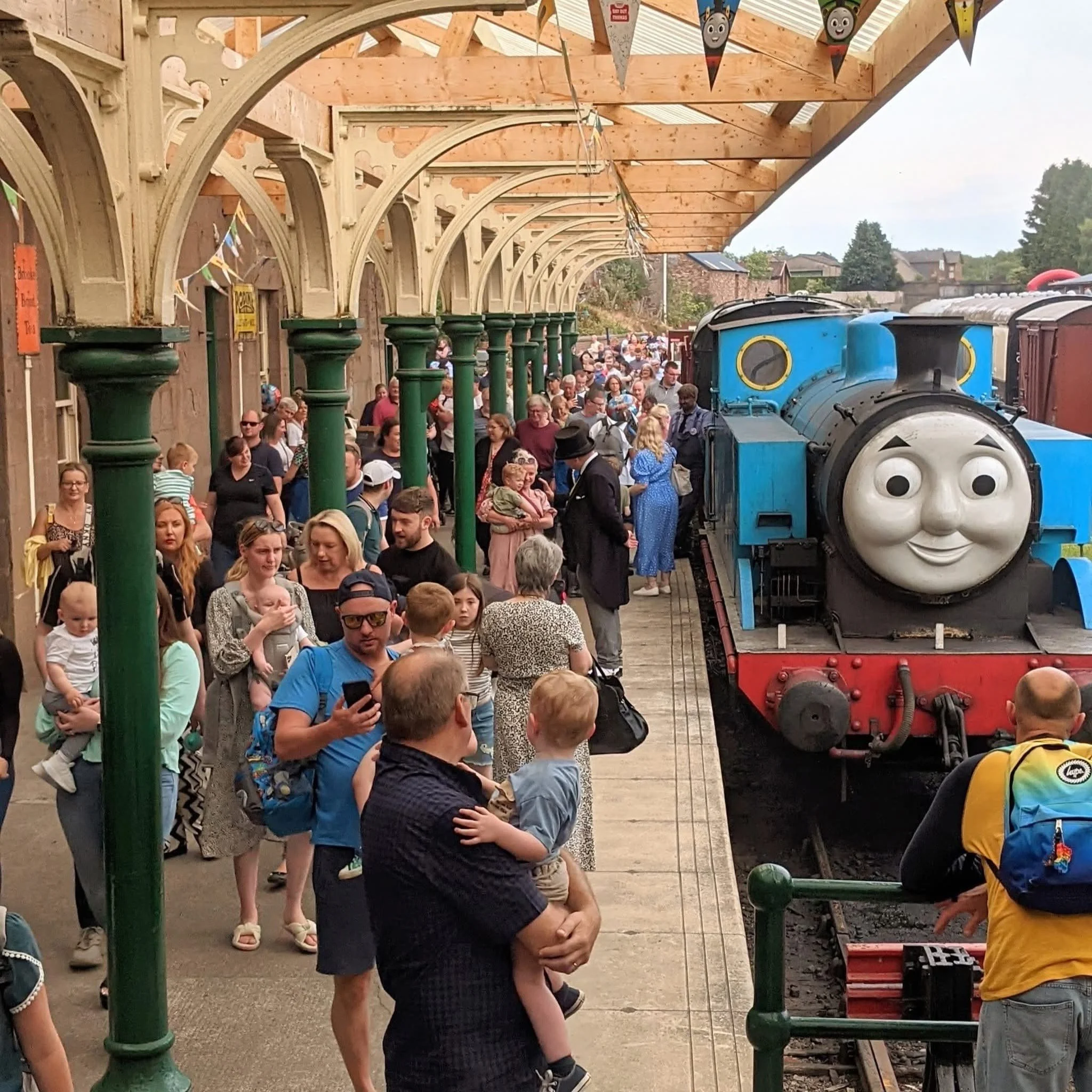 Crowd of people, including children and adults, waiting in line at a train station with a large blue cartoon train and train station shelter with wooden beams and decorative flags.