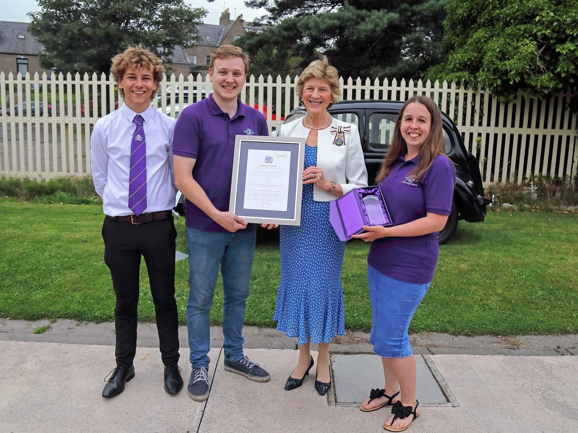 Four volunteers standing together, smiling. The woman in the middle is holding a framed certificate. The girl on the right is holding a purple box celebrating their volunteering achievement.