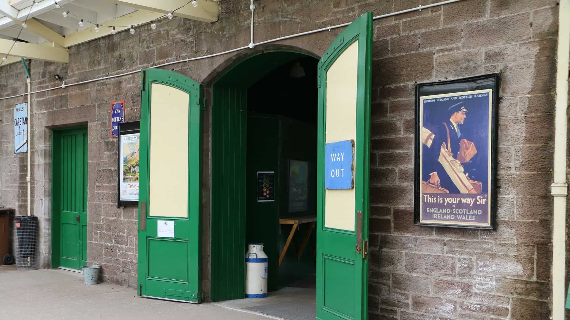 Open green doors of a train station or railway platform with signs and posters on a brick wall. There's a sign that says 'Way Out' and a poster featuring a vintage train conductor with the text 'This is your way Sir'.