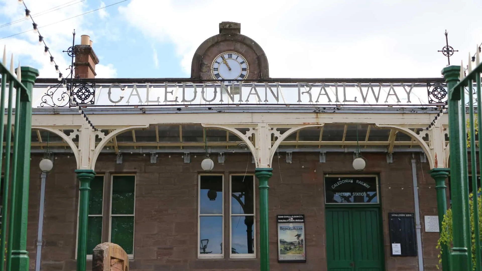 The Caledonian Railway station building with a large clock on top, vintage architecture, green fence in foreground, and sign reading 'Caledonian Railway Brechin Station'.