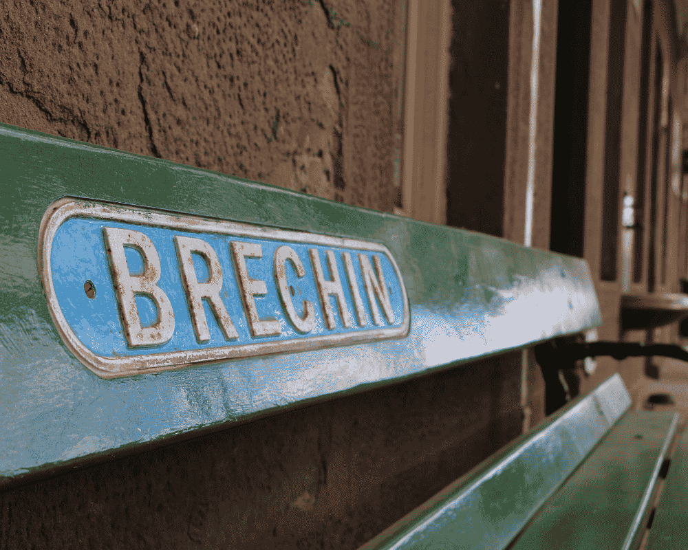 Close-up of a vintage green park bench with a blue and white metal nameplate reading 'Brechin'.