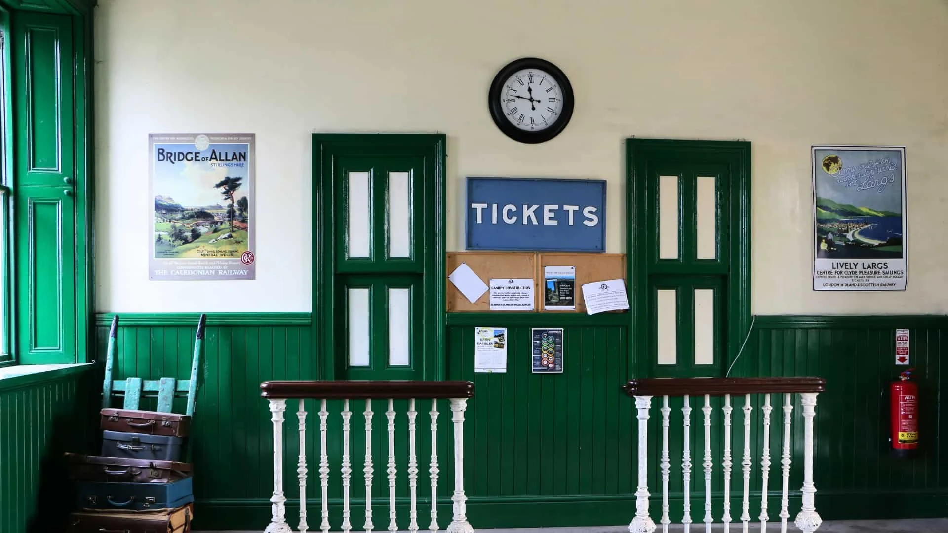 Interior of a ticket office with green paneled doors and wainscoting, a wall clock showing approximately 2:57, and a blue sign with white lettering that says 'TICKETS'. There are posters on the wall, a wooden postal rack with vintage suitcases underneath, and a red fire extinguisher in the corner.