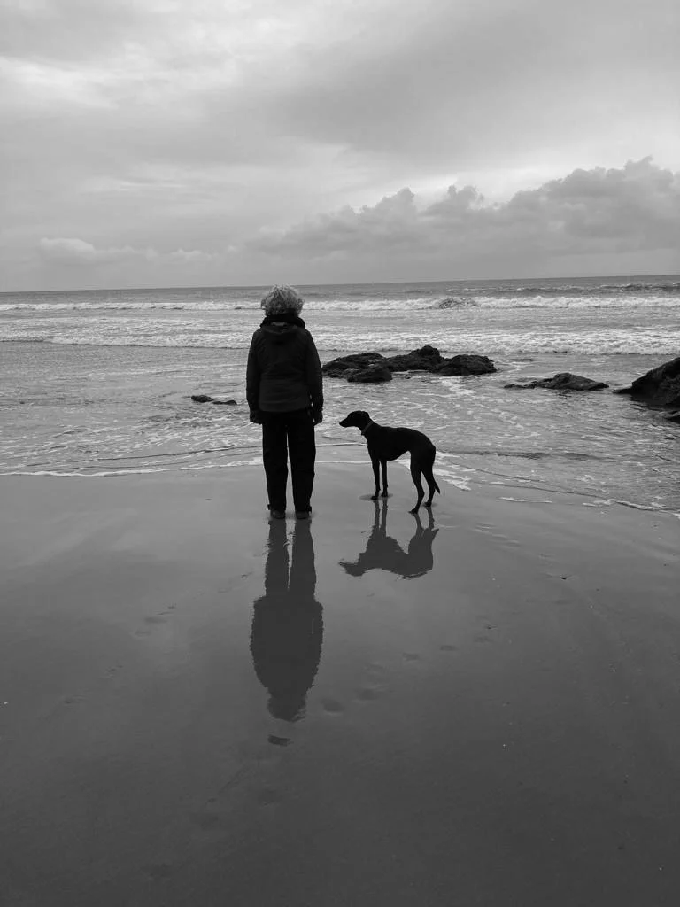 A person standing on a beach with a dog, looking out at the ocean with cloudy sky, black and white photography.