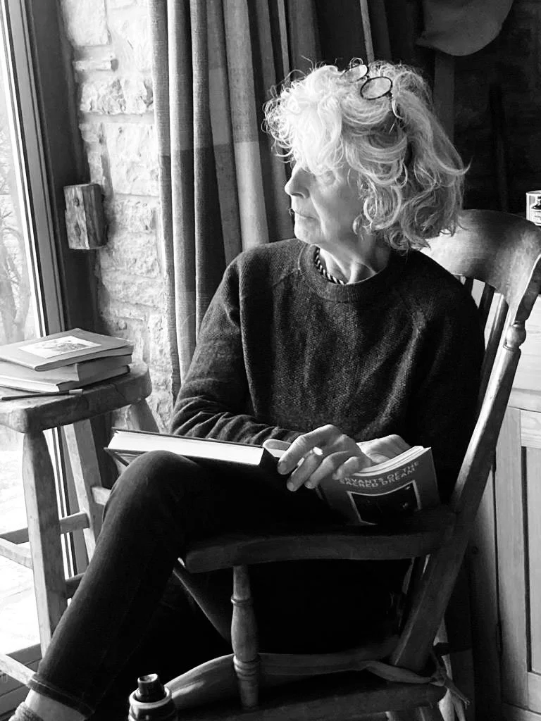 A woman with curly, shoulder-length hair and glasses resting on her head, sitting in a wooden chair, looking out a window, holding a book, with a stack of books on a small table nearby.