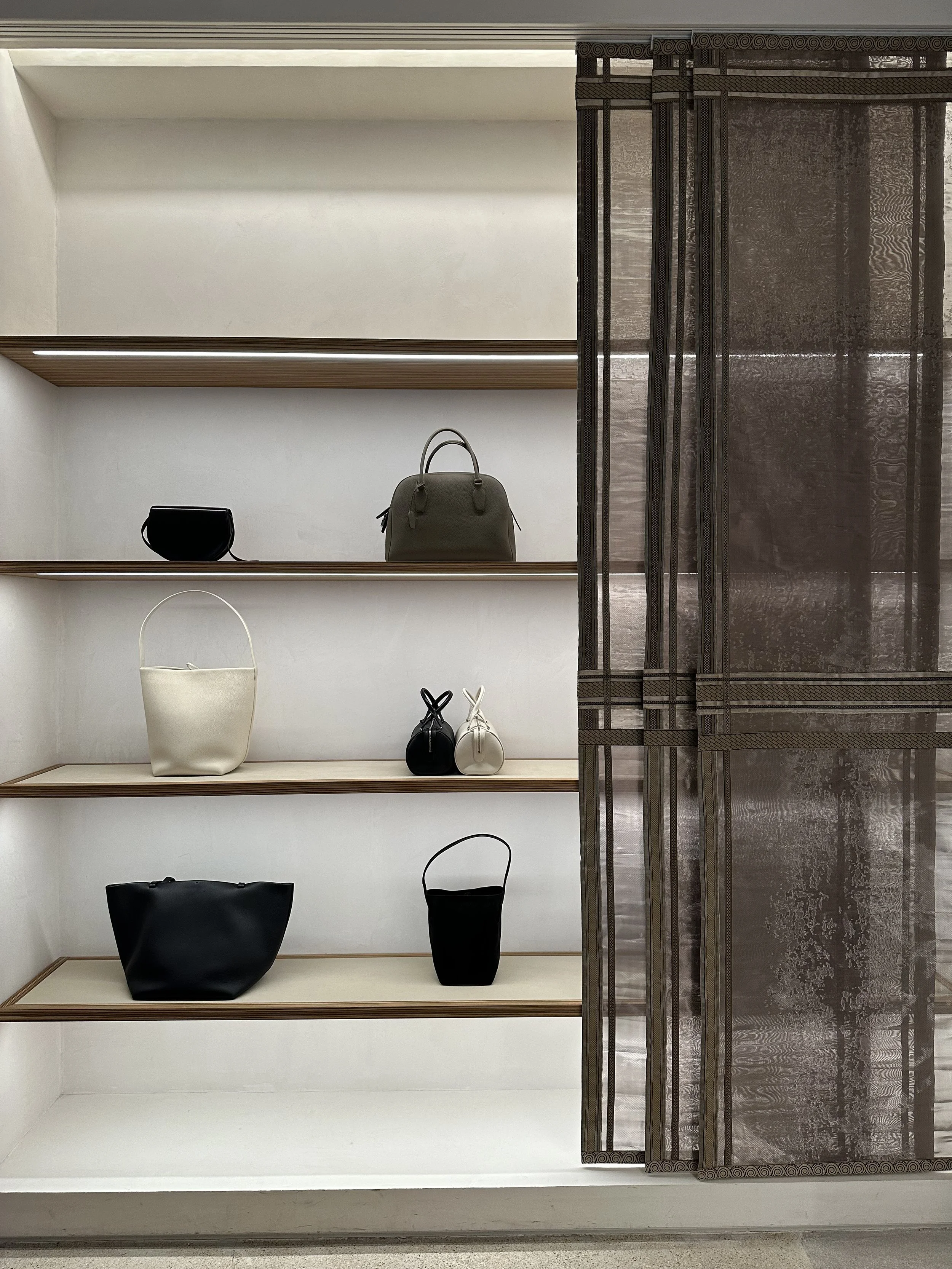 Display of handbags on white shelves behind a sheer curtain in a retail store.