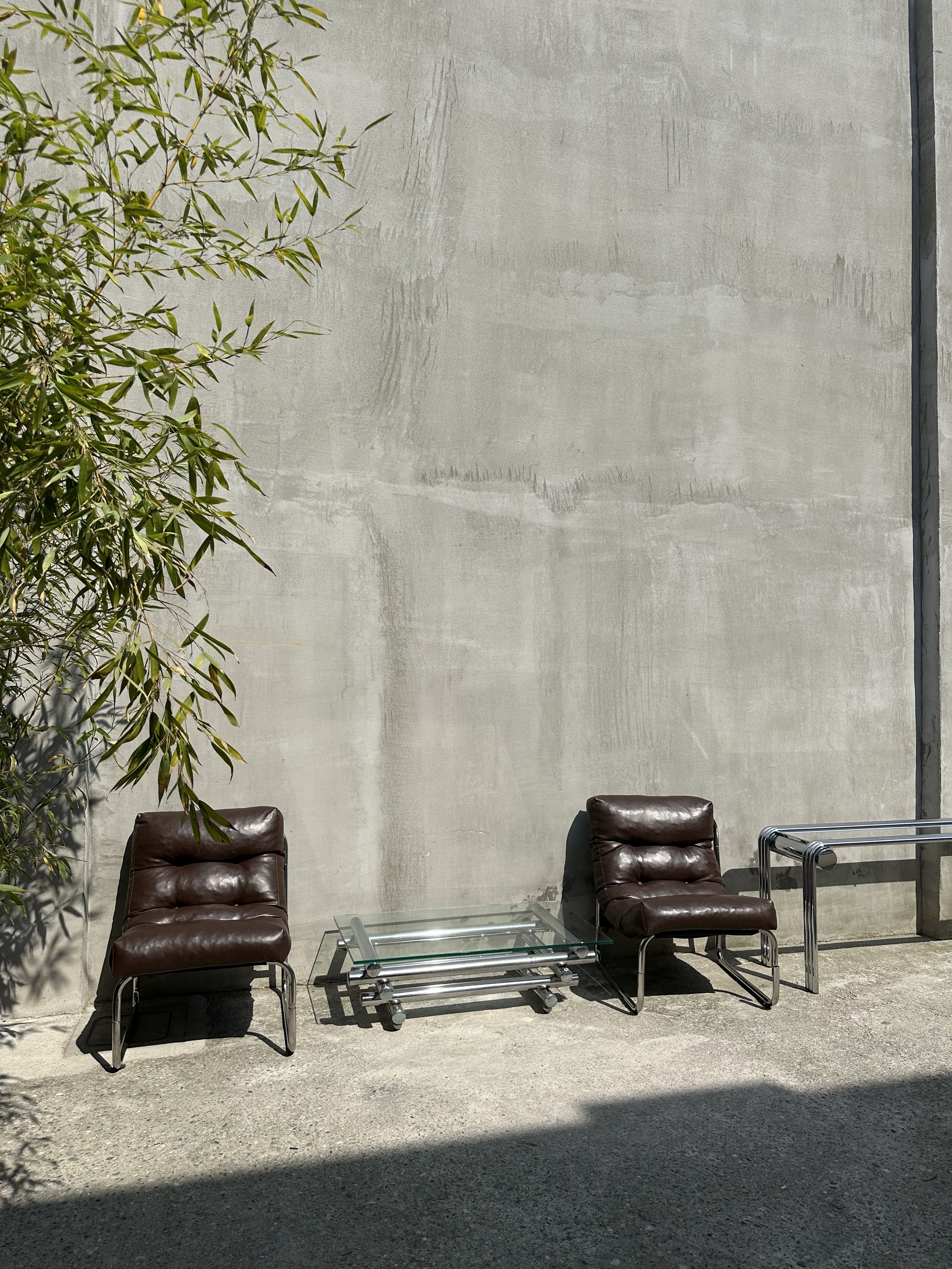 Two brown leather lounge chairs and a glass coffee table placed outside against a plain concrete wall, with a tree on the left side casting shadows.