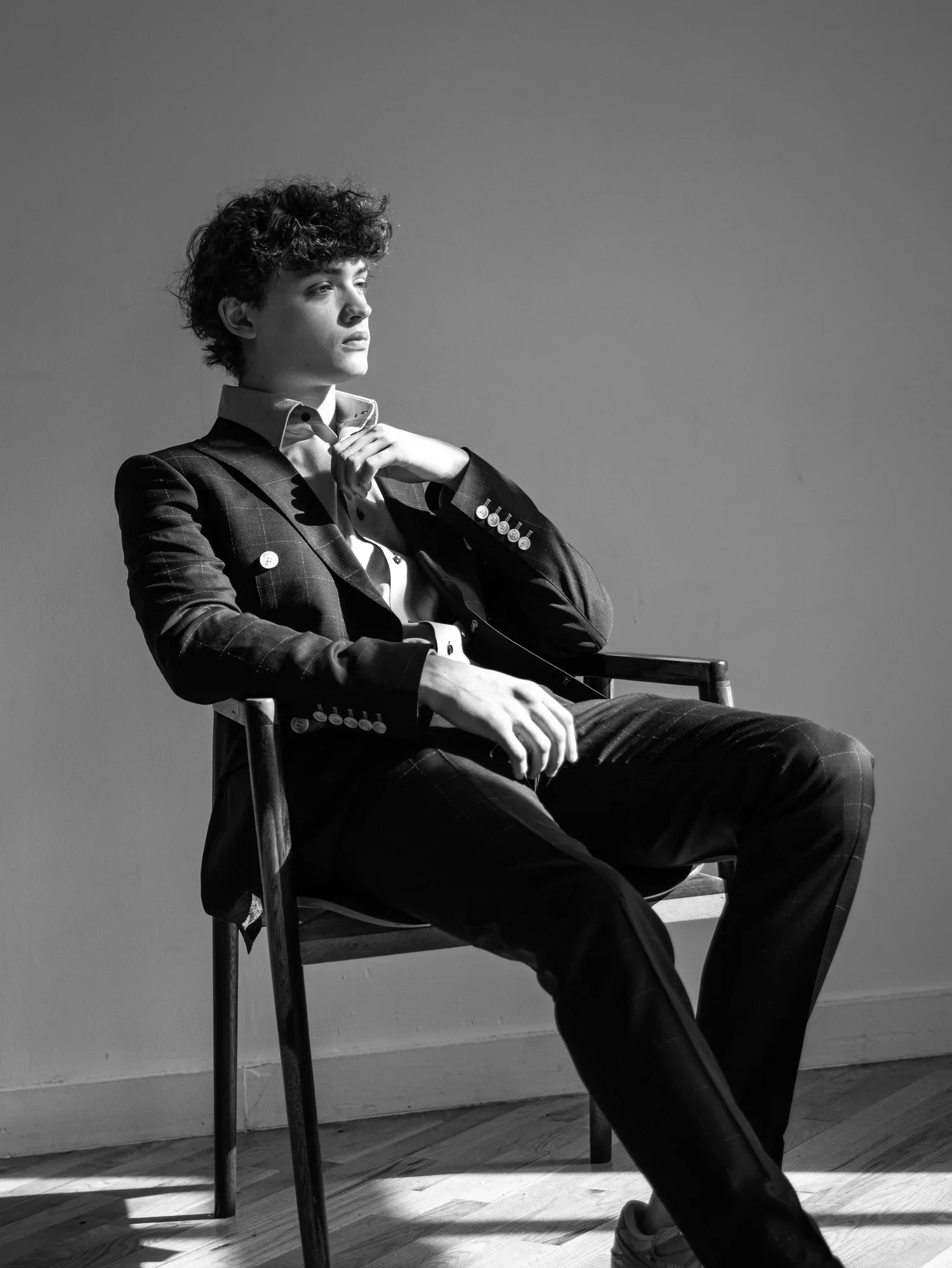 A black and white photo of a young person sitting on a chair, looking to the side. They are wearing a suit and have curly hair.