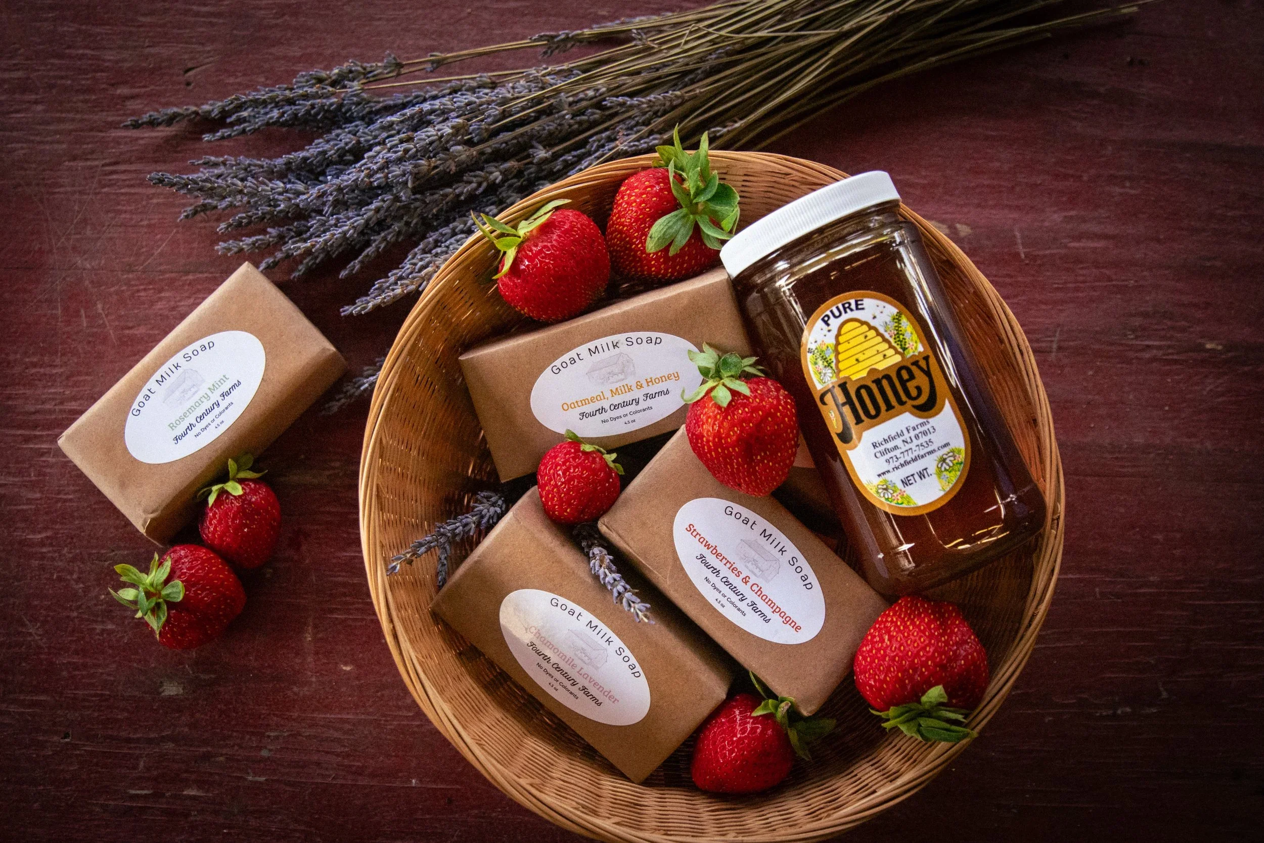 Basket containing homemade goat milk soaps with strawberry, lavender, and honey scent labels, fresh strawberries, and a jar of honey, on a dark wooden surface with dried lavender.